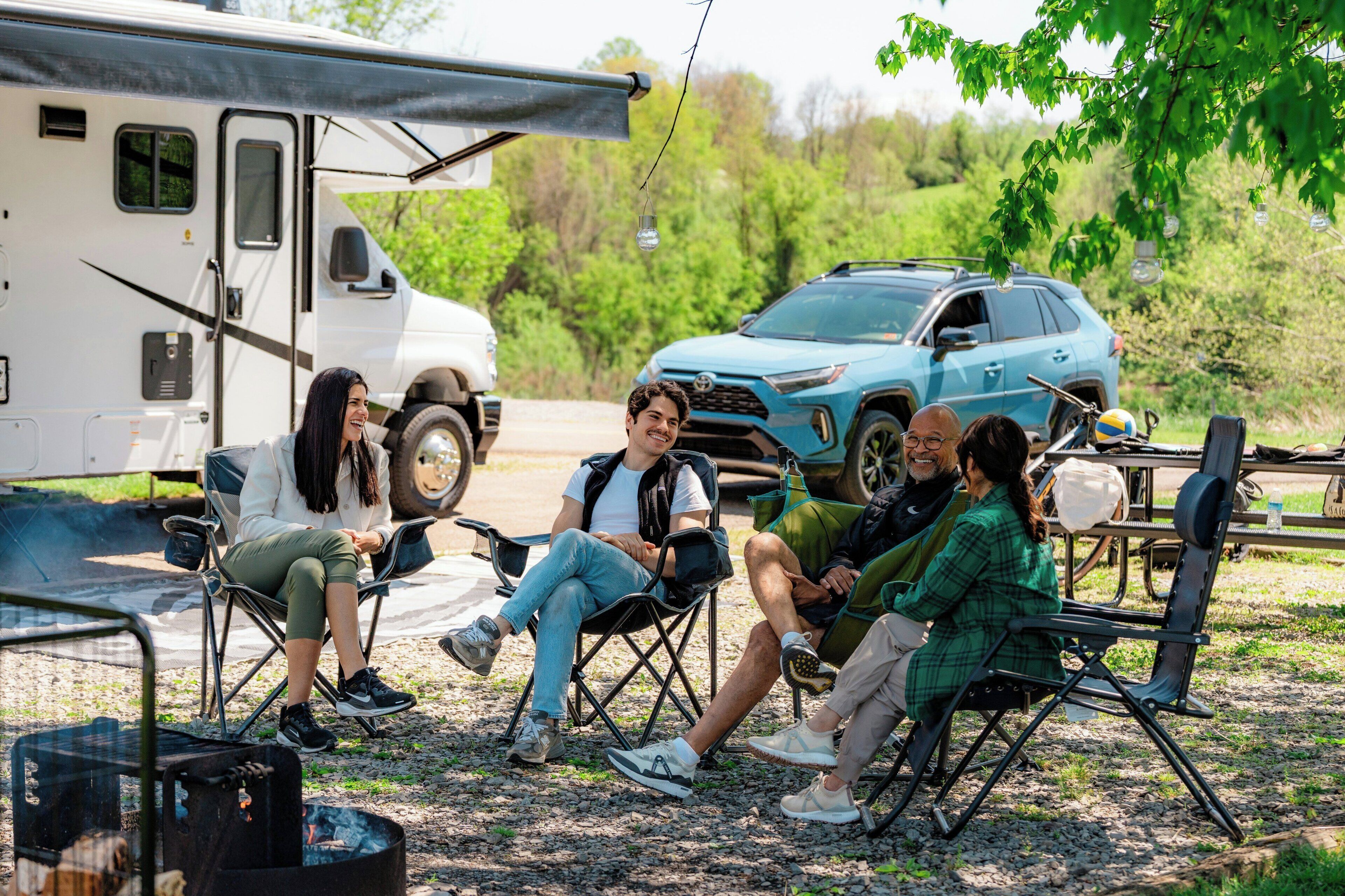 A group of people sitting on camping chairs outdoors with a vehicle and a tent in the background.