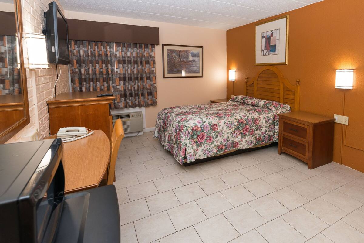 A hotel room with a bed, bedside tables, a desk, a chair, a TV, a mirror, a window with curtains, and a wall-mounted picture.