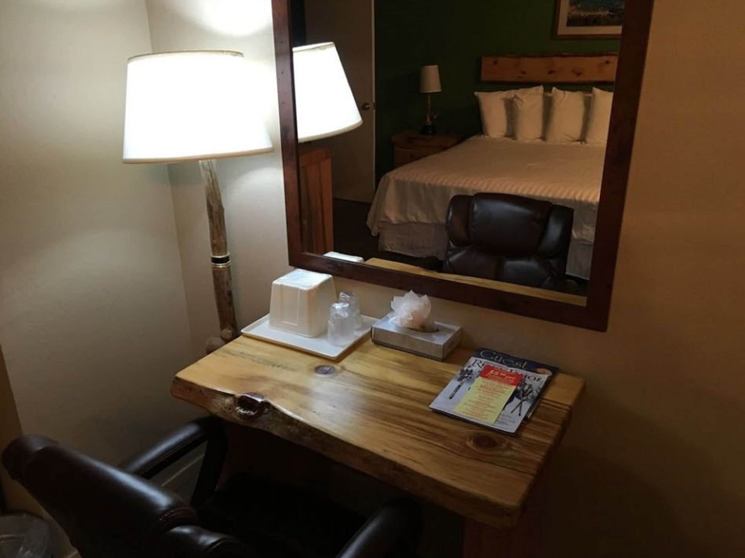A hotel room with a wooden desk, two lamps, a mirror, a bed, a chair, and a magazine.