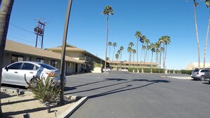 A street view with a row of palm trees, parked cars, and a building with a balcony on the left. - Americas Best Value Inn Blythe (Blythe)