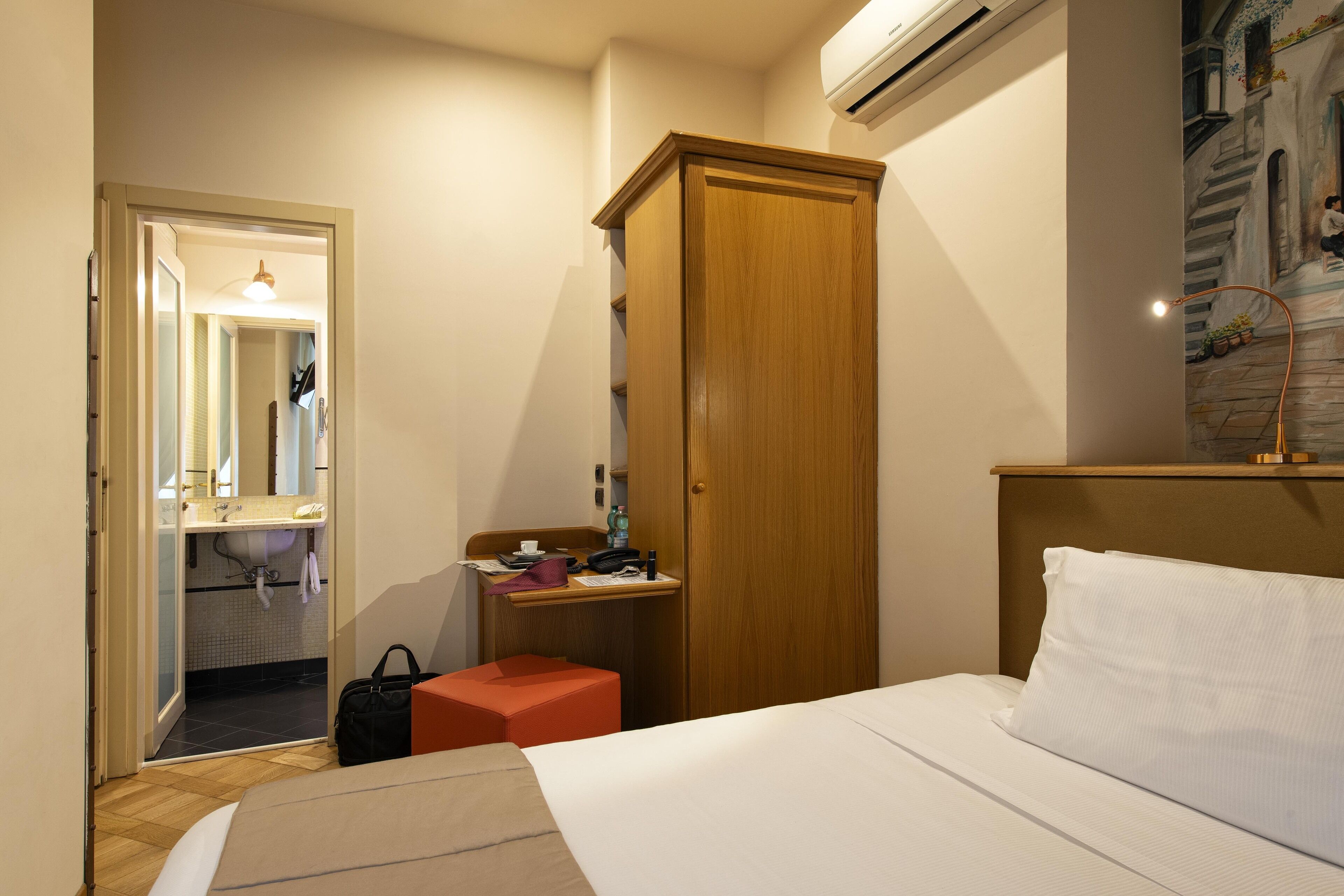 A hotel room with a bed, a wooden wardrobe, a small desk with a chair, and a bathroom visible through an open door.