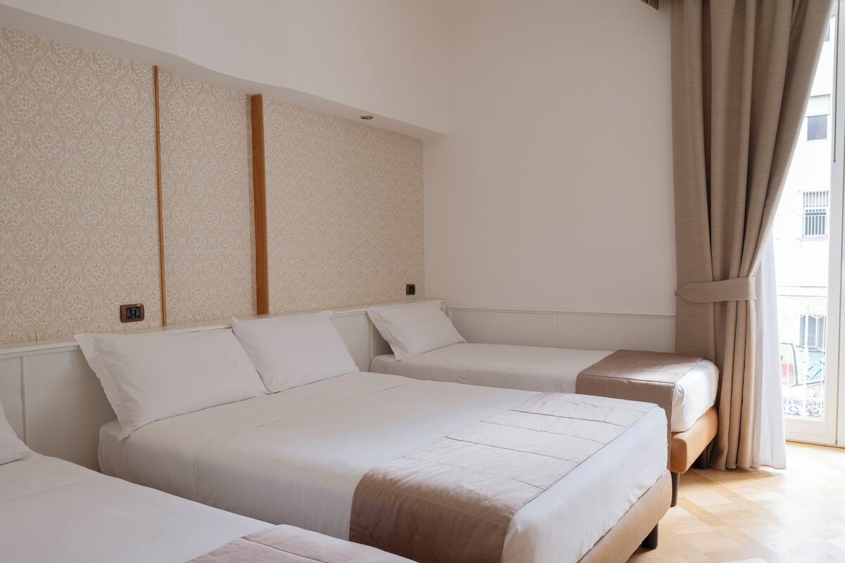 A hotel room with three beds arranged in a row, a large window with curtains, and a patterned wallpaper on the walls.