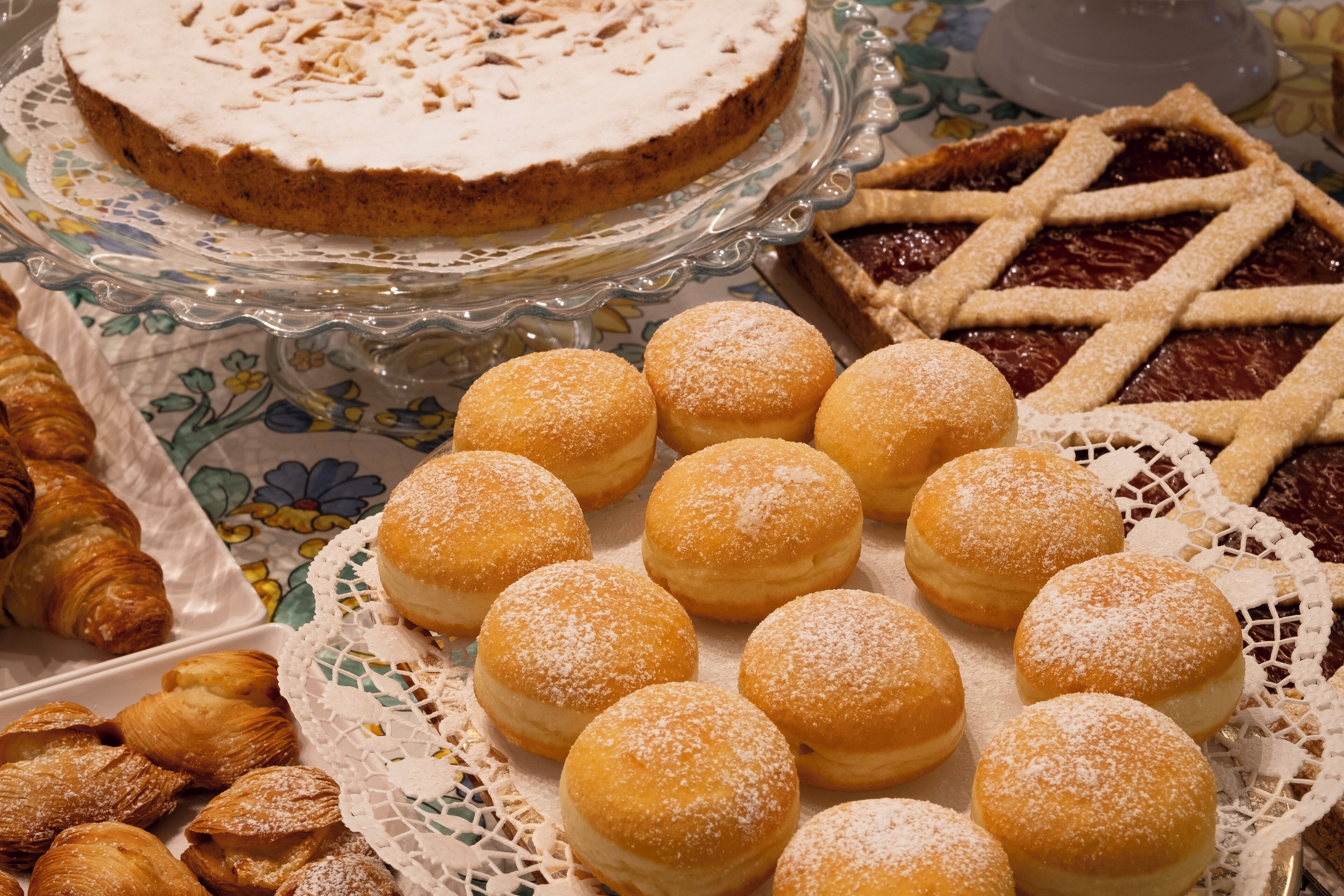 A variety of pastries including a round cake with a lattice crust, a pie with a lattice crust, and several round pastries dusted with powdered sugar.