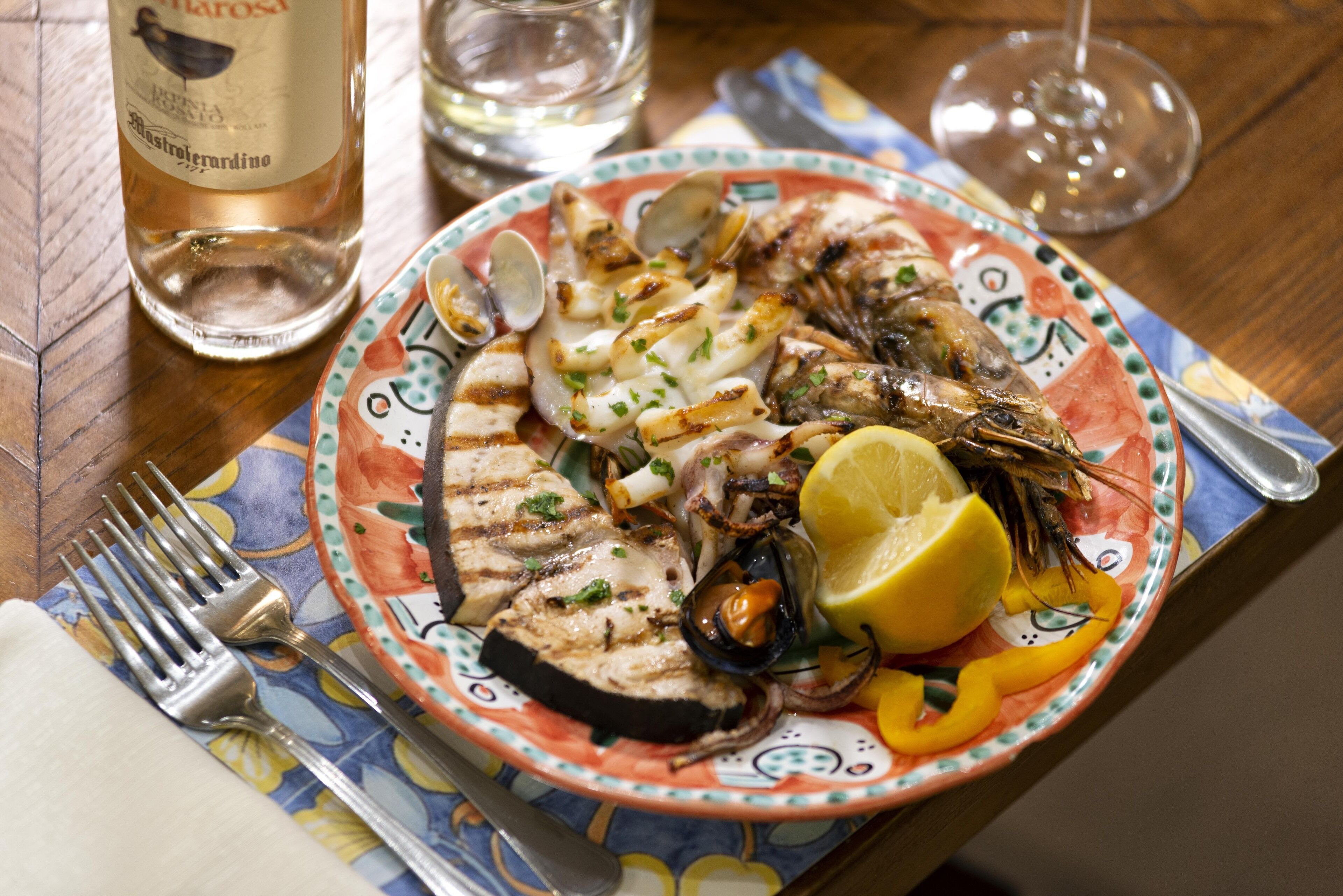 Une assiette de fruits de mer grillés, arrosés de citron et parsemés d’herbes, une bouteille de vin Astroturardino et un verre d’eau sur une table.