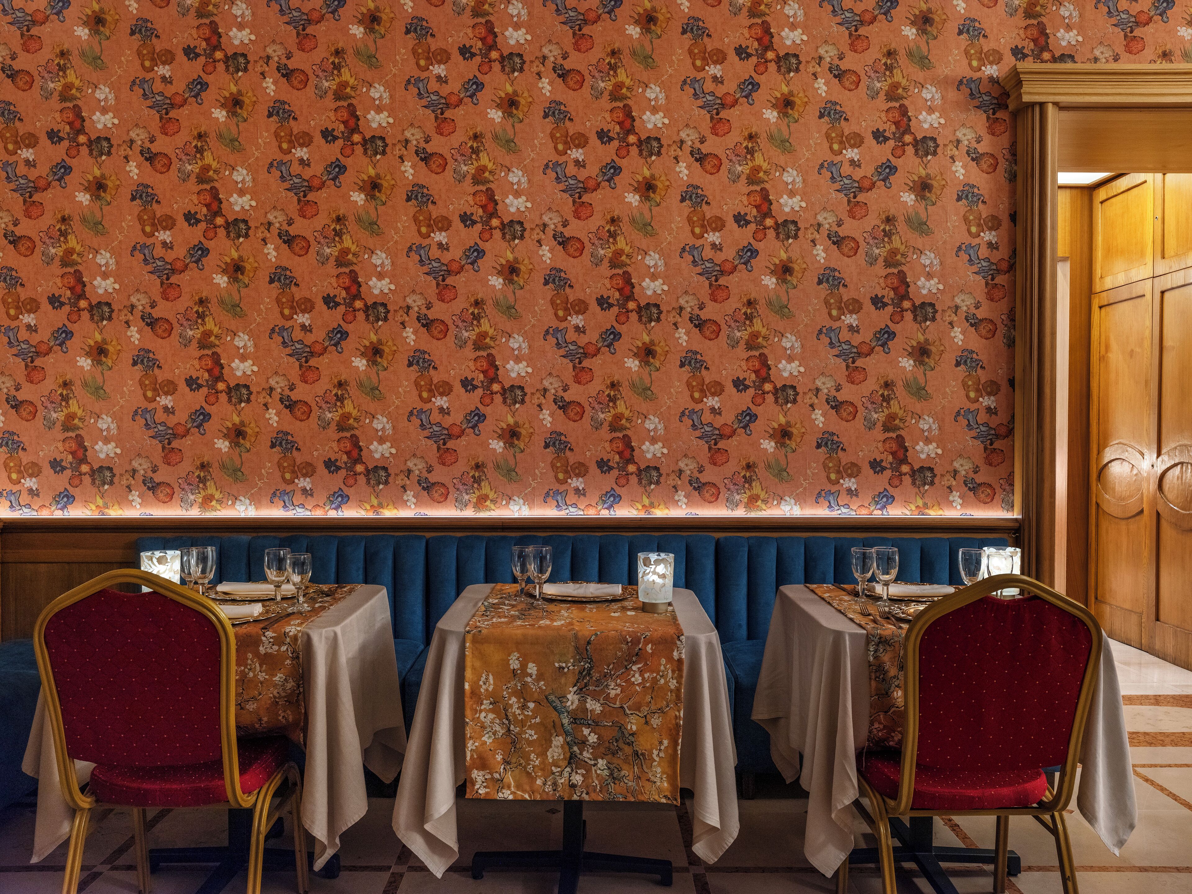 A restaurant with three tables, each with a white tablecloth and red chairs. The wall behind the tables is covered with a floral wallpaper.
