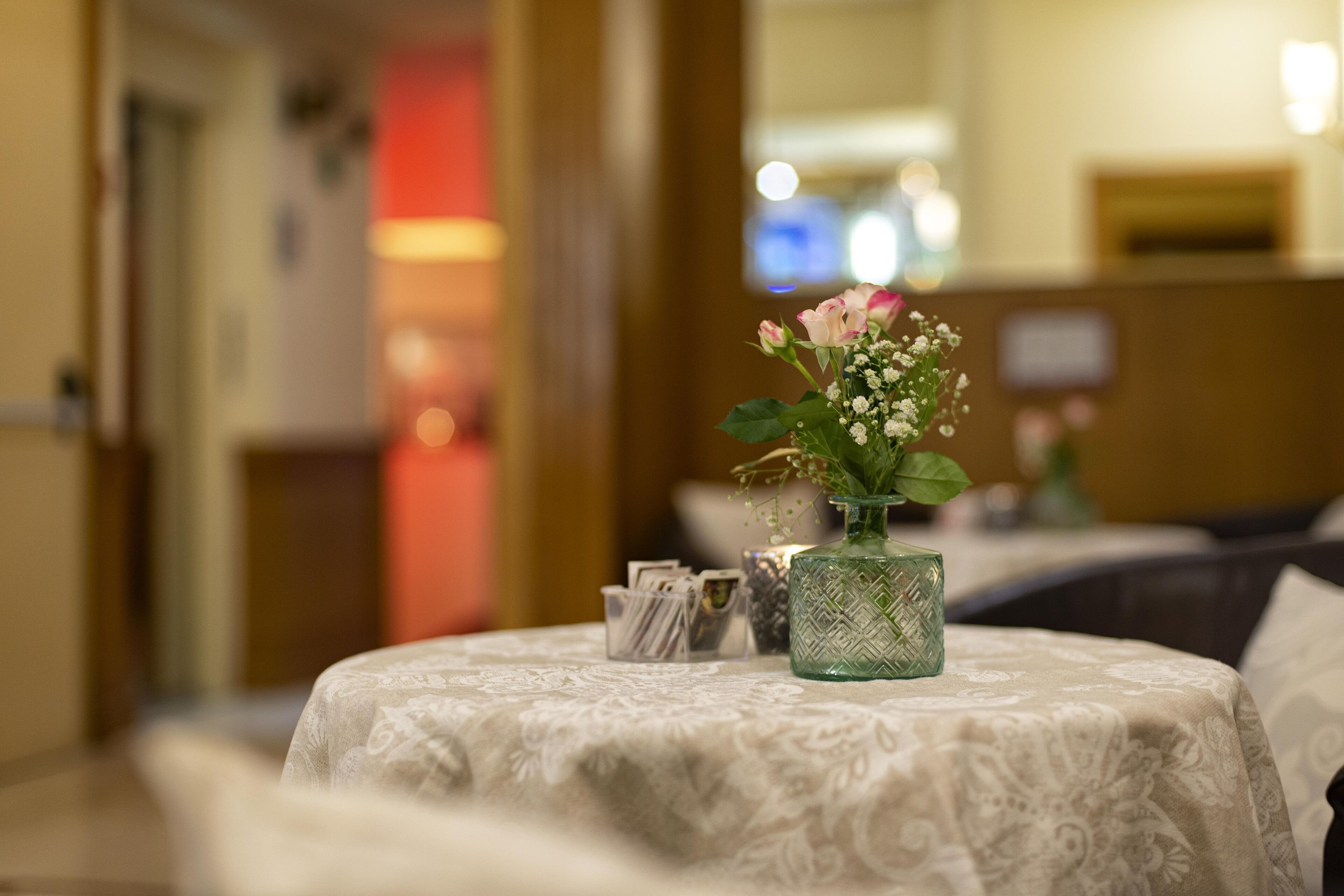 Une chambre d’hôtel avec une table ronde recouverte d’une nappe à motifs, un vase avec des fleurs et un petit objet décoratif posé sur la table.