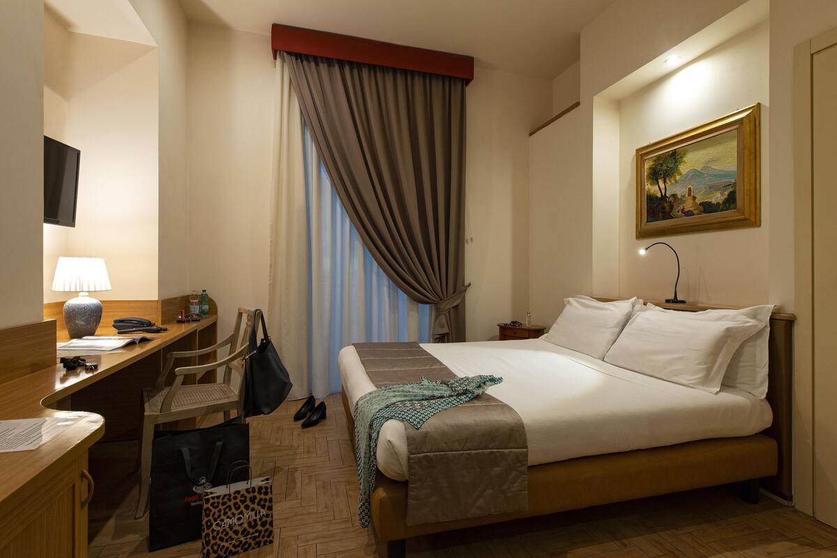 A hotel room with a bed, desk, chair, and a painting on the wall.
