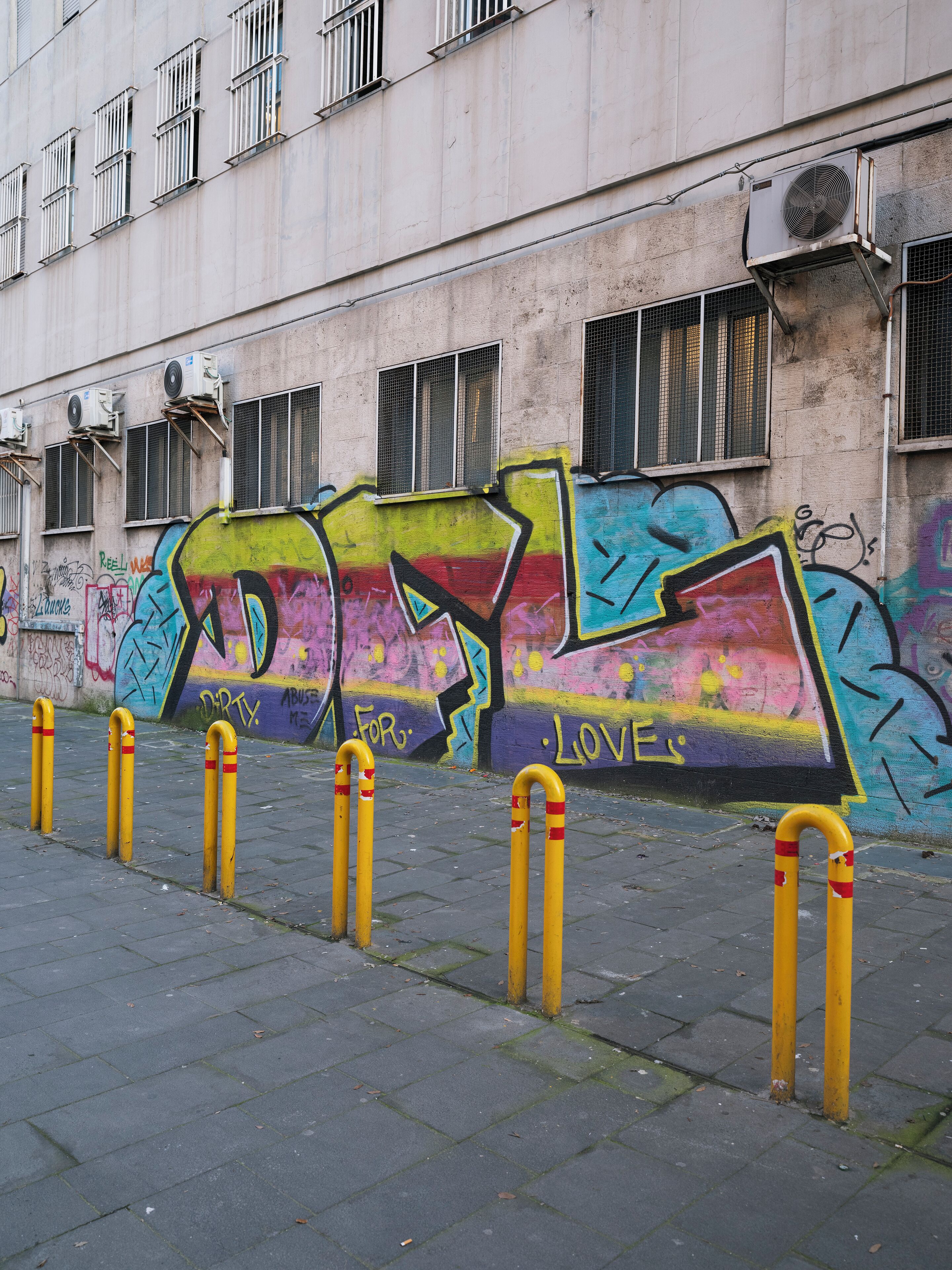 Fachada de edificio con grafitis, unidades de aire acondicionado, ventanas con barrotes y bolardos amarillos en una acera pavimentada.