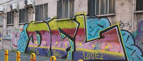 A building facade with graffiti, air conditioning units, barred windows, and yellow bollards on a paved sidewalk.