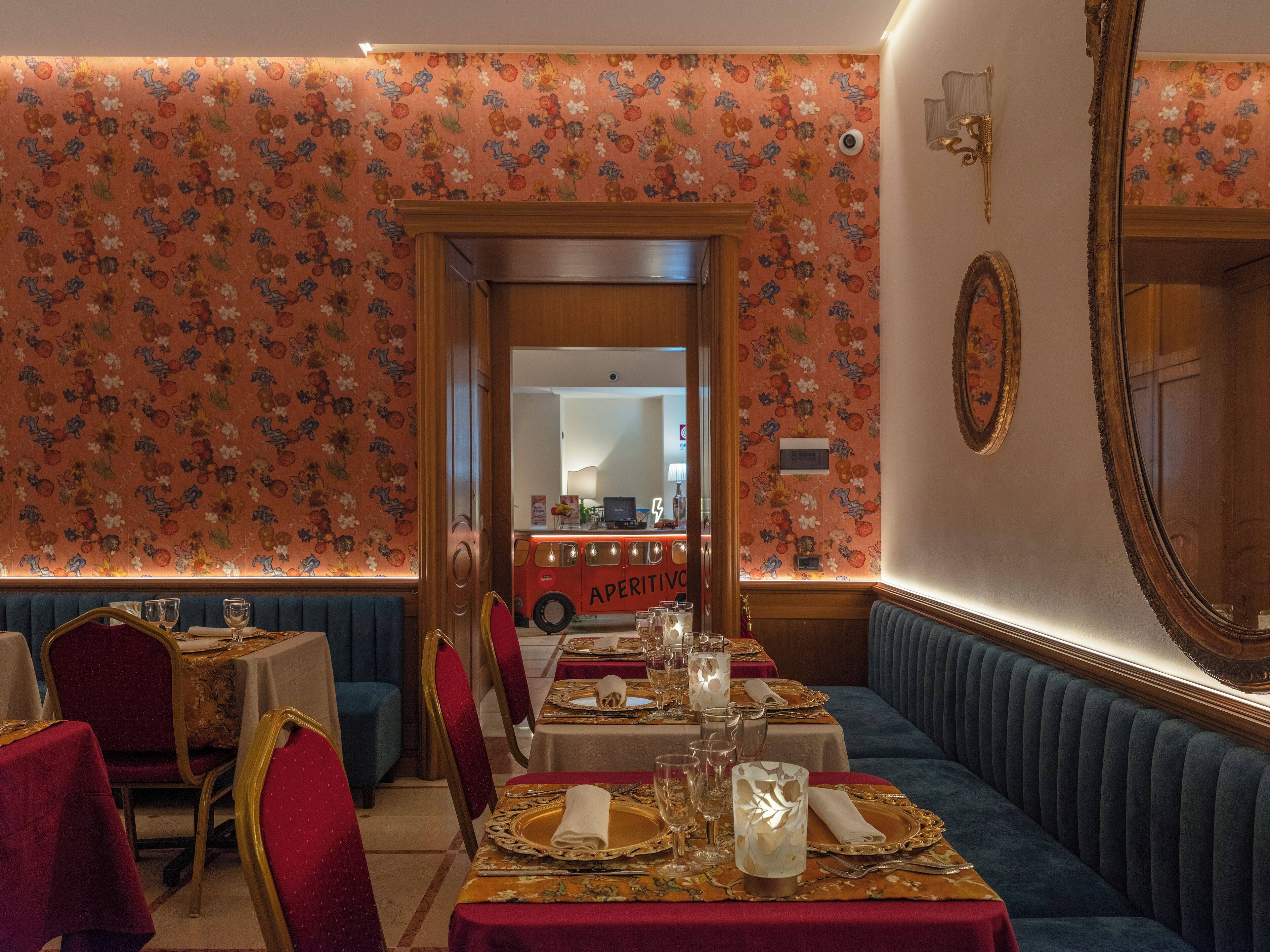 A restaurant with floral wallpaper, red and gold table settings, and a vintage red bus in the background.
