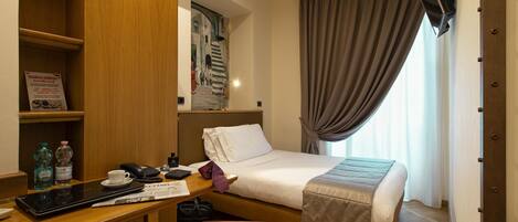 A hotel room with a bed, a desk with a computer, a chair, a small table with a cup and a bottle, and a window with curtains.