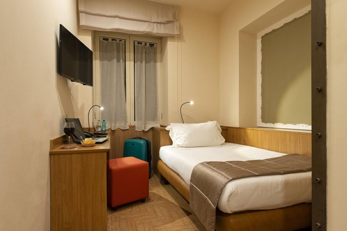 A hotel room with a bed, a desk with a computer, a TV, a window with curtains, and a suitcase.