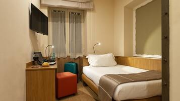 A hotel room with a bed, a desk with a computer, a TV, a window with curtains, and a suitcase.