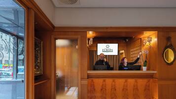 Een hotel receptie met een houten balie, een grote spiegel en een bord met daarop Grand Hotel Europa in Napels.