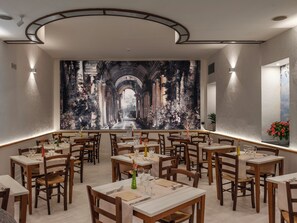 A restaurant with multiple tables and chairs, a large mural on the wall, and a view of the outdoors. - Grand Hotel Europa (Naples)