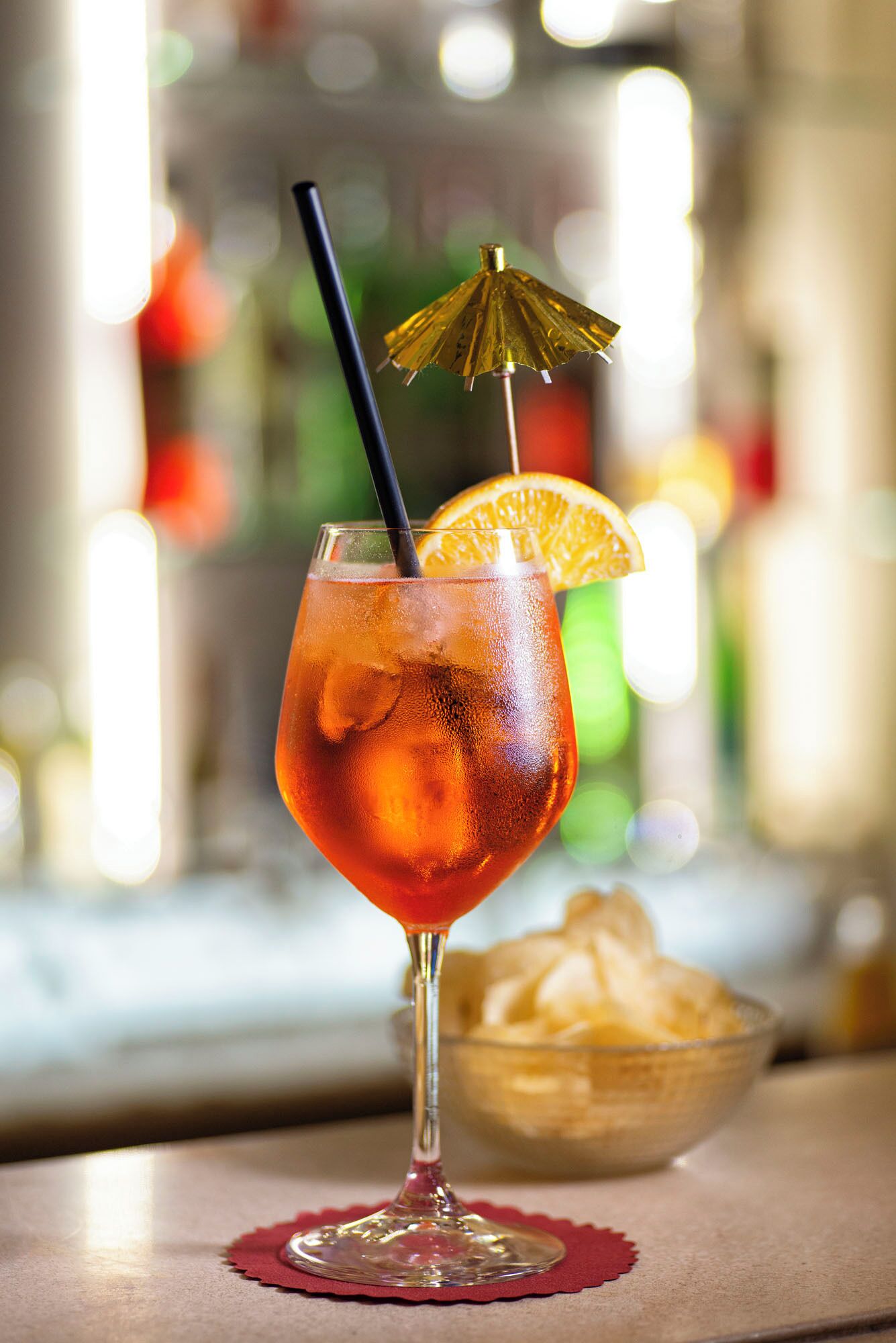 Un verre à cocktail avec une paille, une tranche d’orange et un petit parapluie doré, le tout posé sur un sous-verre rouge.