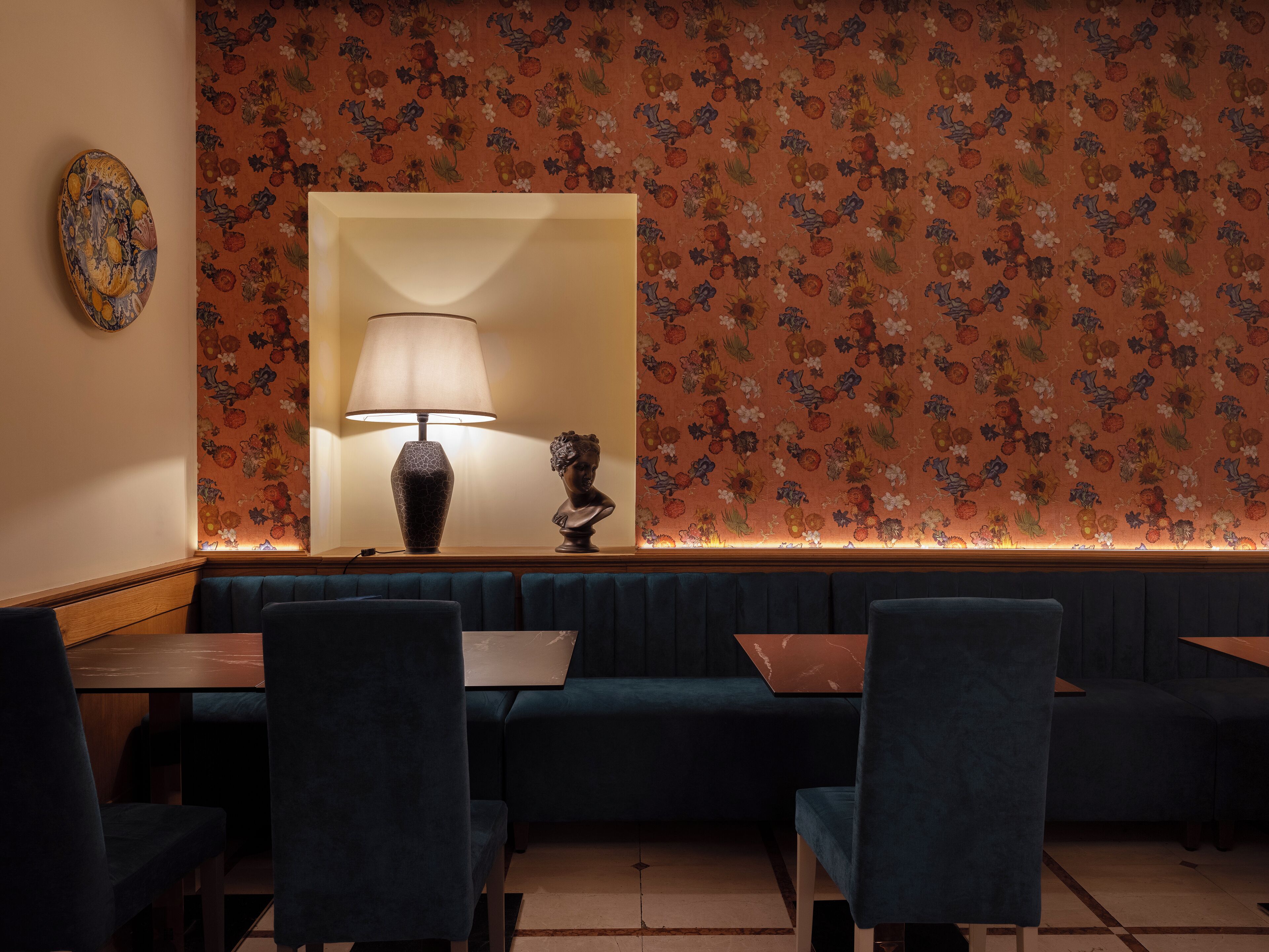 A restaurant interior with a floral wallpaper, a table lamp, a bust sculpture, and blue upholstered seating.
