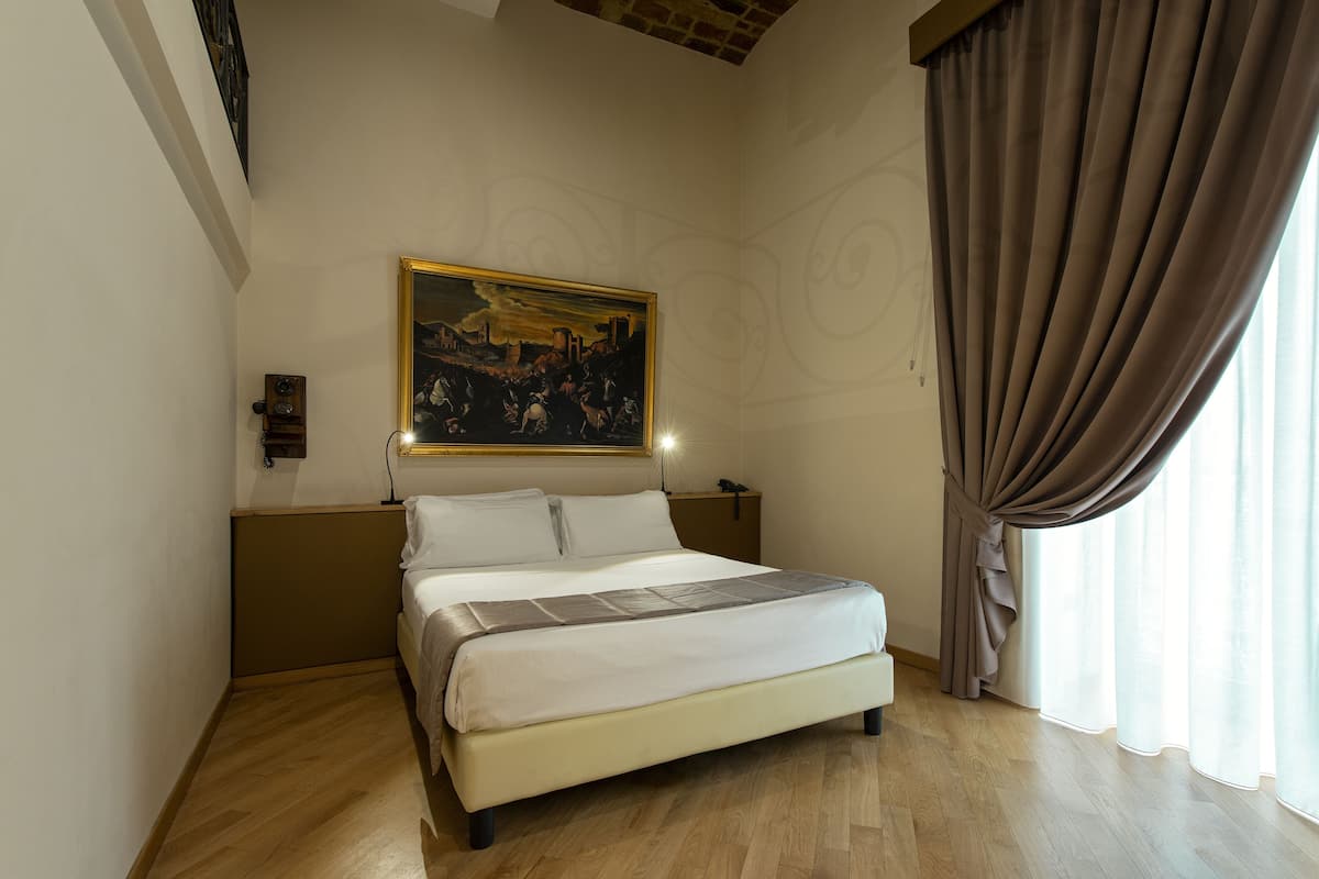 A neatly made bed with a white comforter and a gray blanket, a painting of a cityscape on the wall, and a large window with curtains.