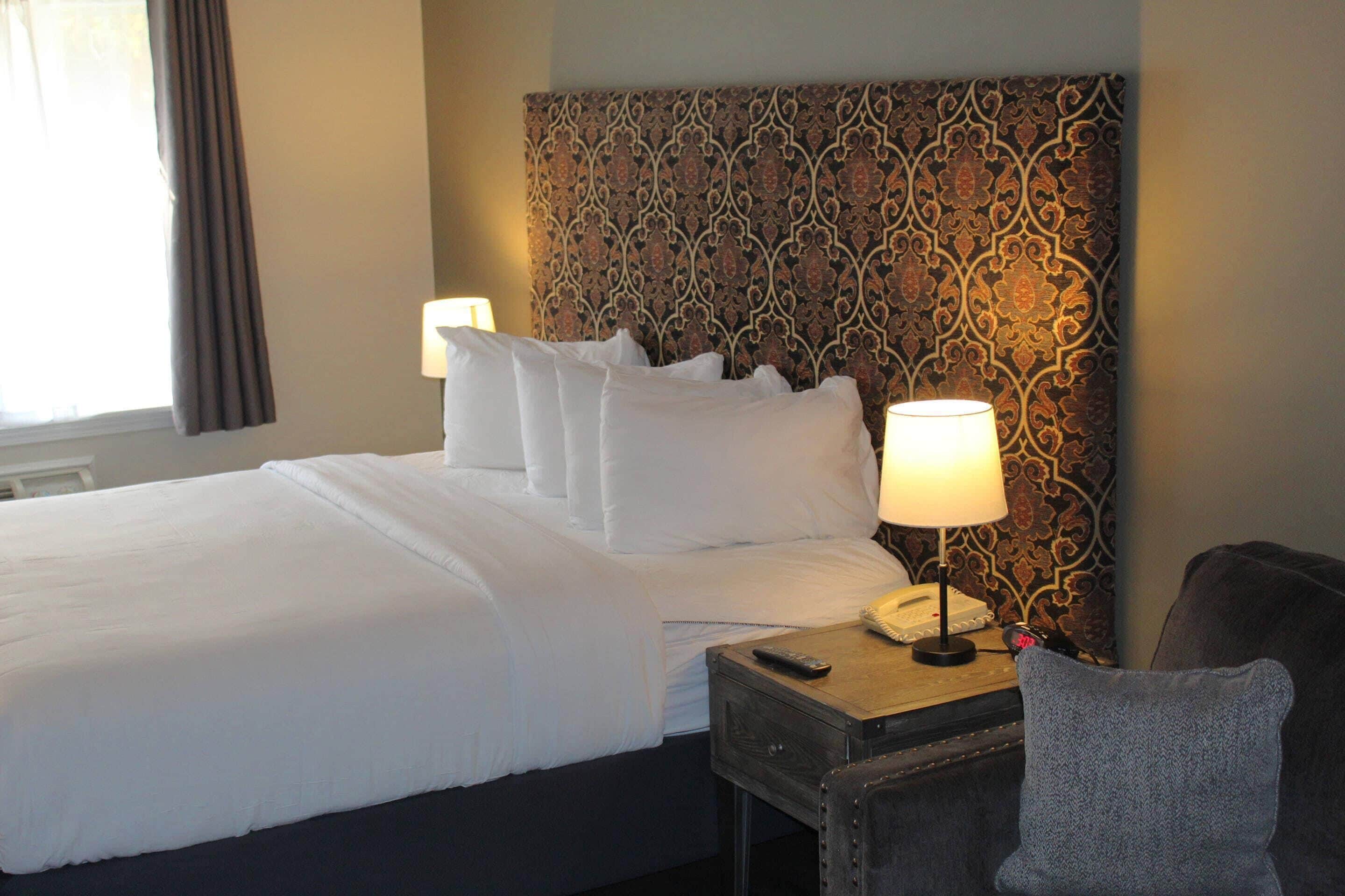 Quarto de hotel com cama bem arrumada, mesa de cabeceira com abajur e telefone, e uma cadeira.