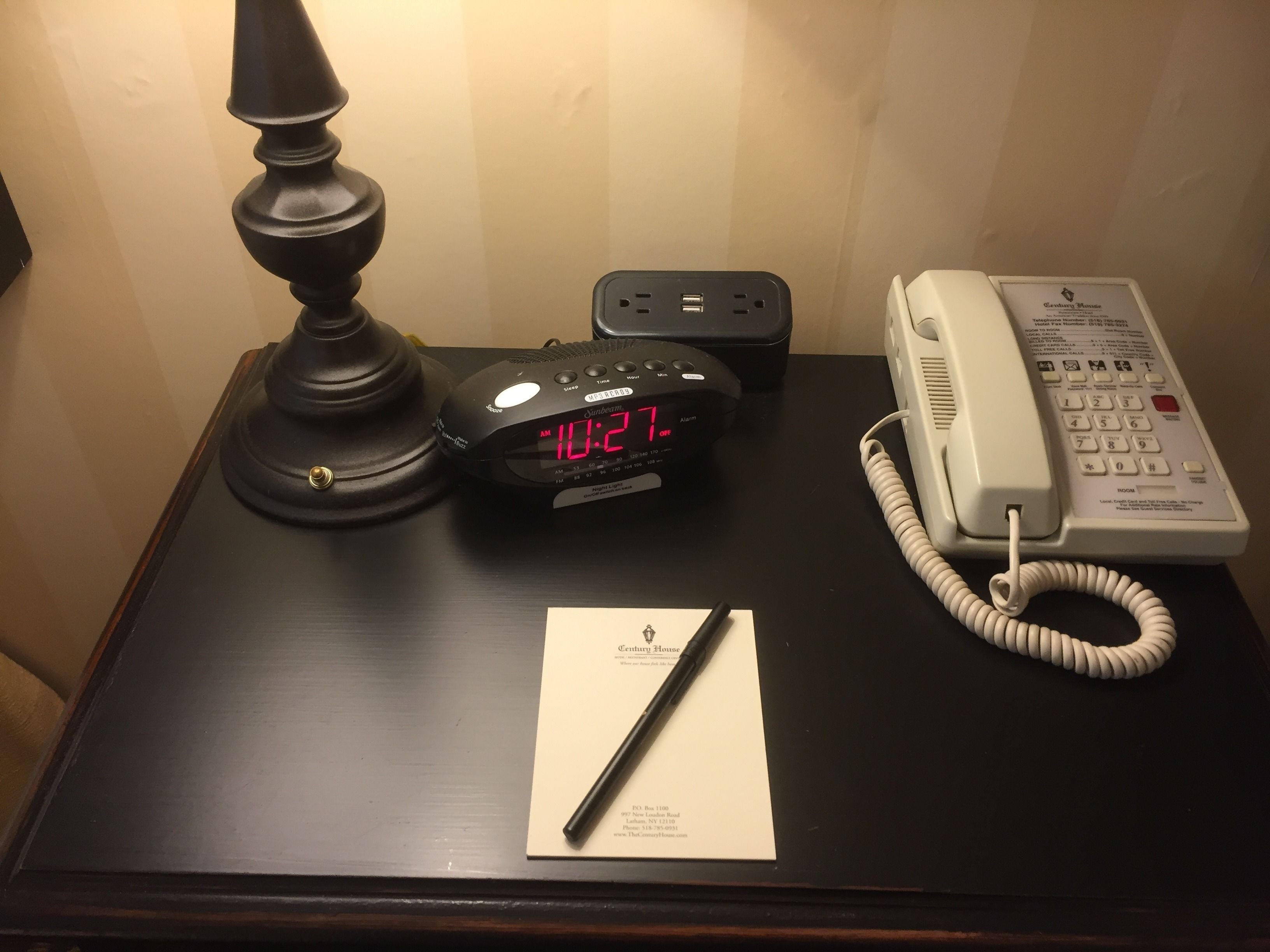 A wooden nightstand with a black lamp, a digital clock displaying 10:17 AM, a white telephone, and a notepad with a pen.