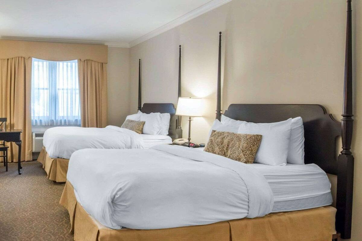 A hotel room with two beds, beige curtains, a desk, and a lamp.
