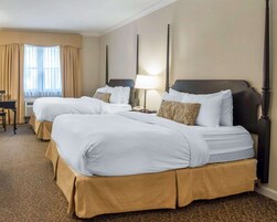 A hotel room with two beds, beige curtains, a desk, and a lamp.