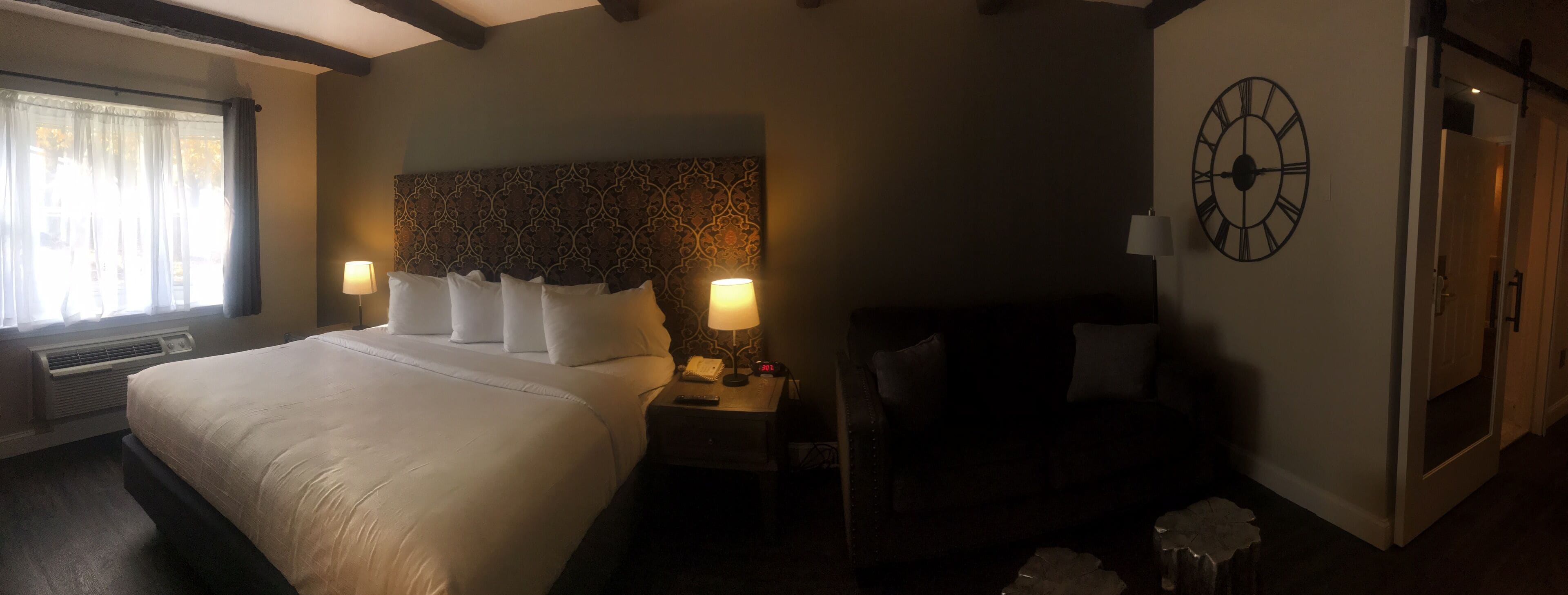 A hotel room with a large bed, two bedside lamps, a clock on the wall, and a window with curtains.