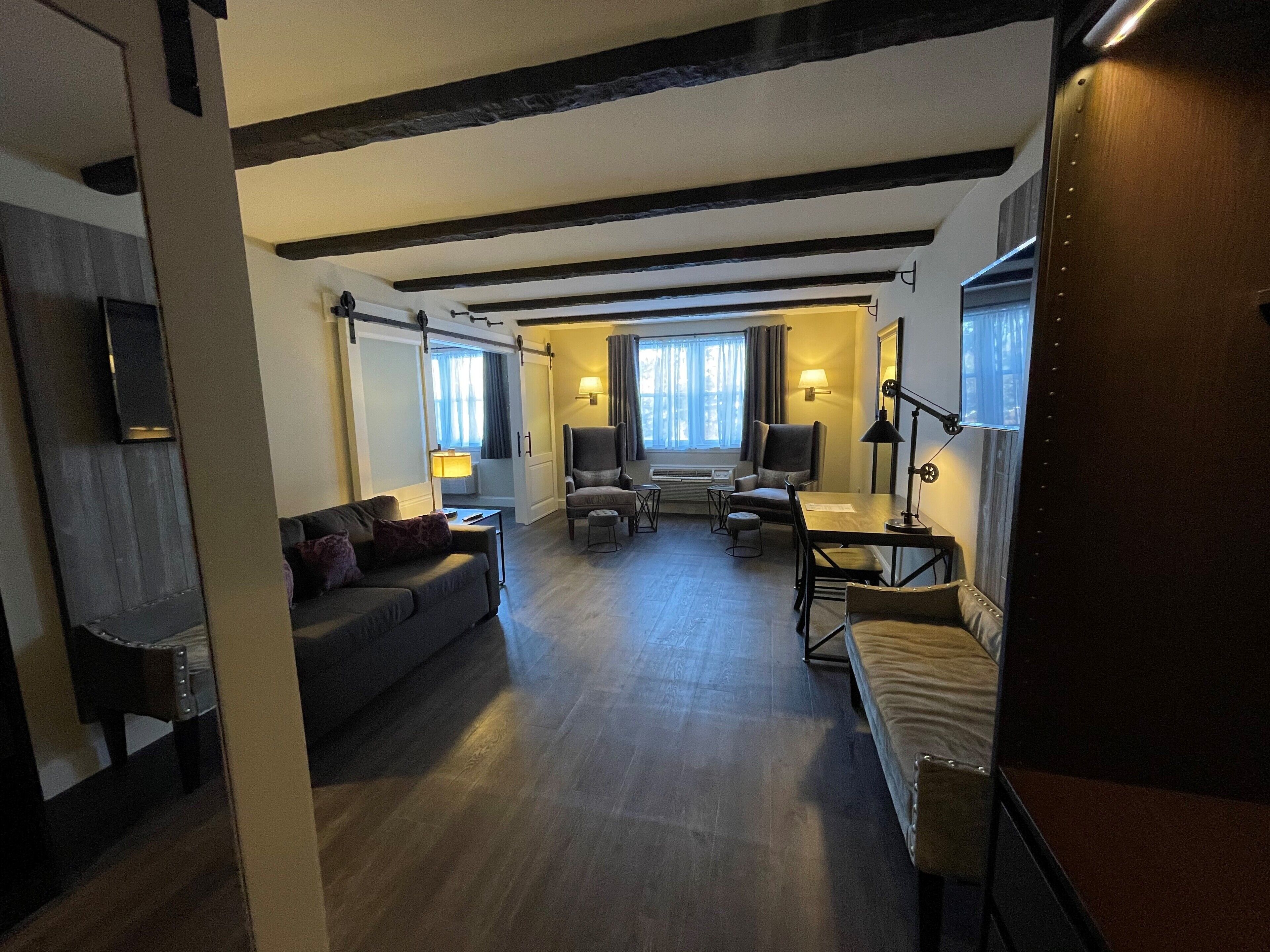 A hotel room with a dark wooden ceiling, a sofa, a desk with a chair, and a window with curtains.