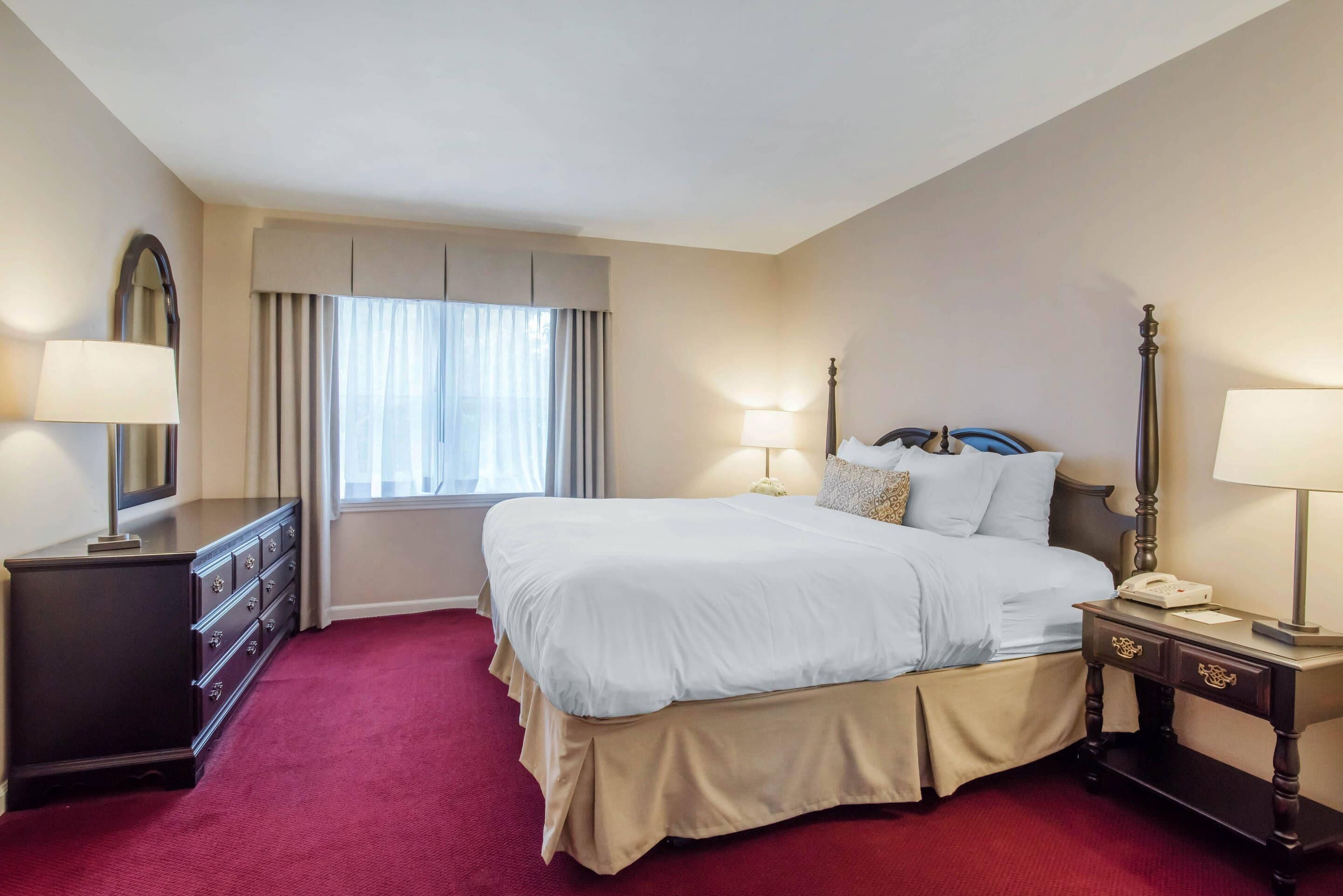 Ein Hotelzimmer mit einem großen Bett, einem Schminktisch, einem Nachttisch und einem Fenster mit Vorhängen.