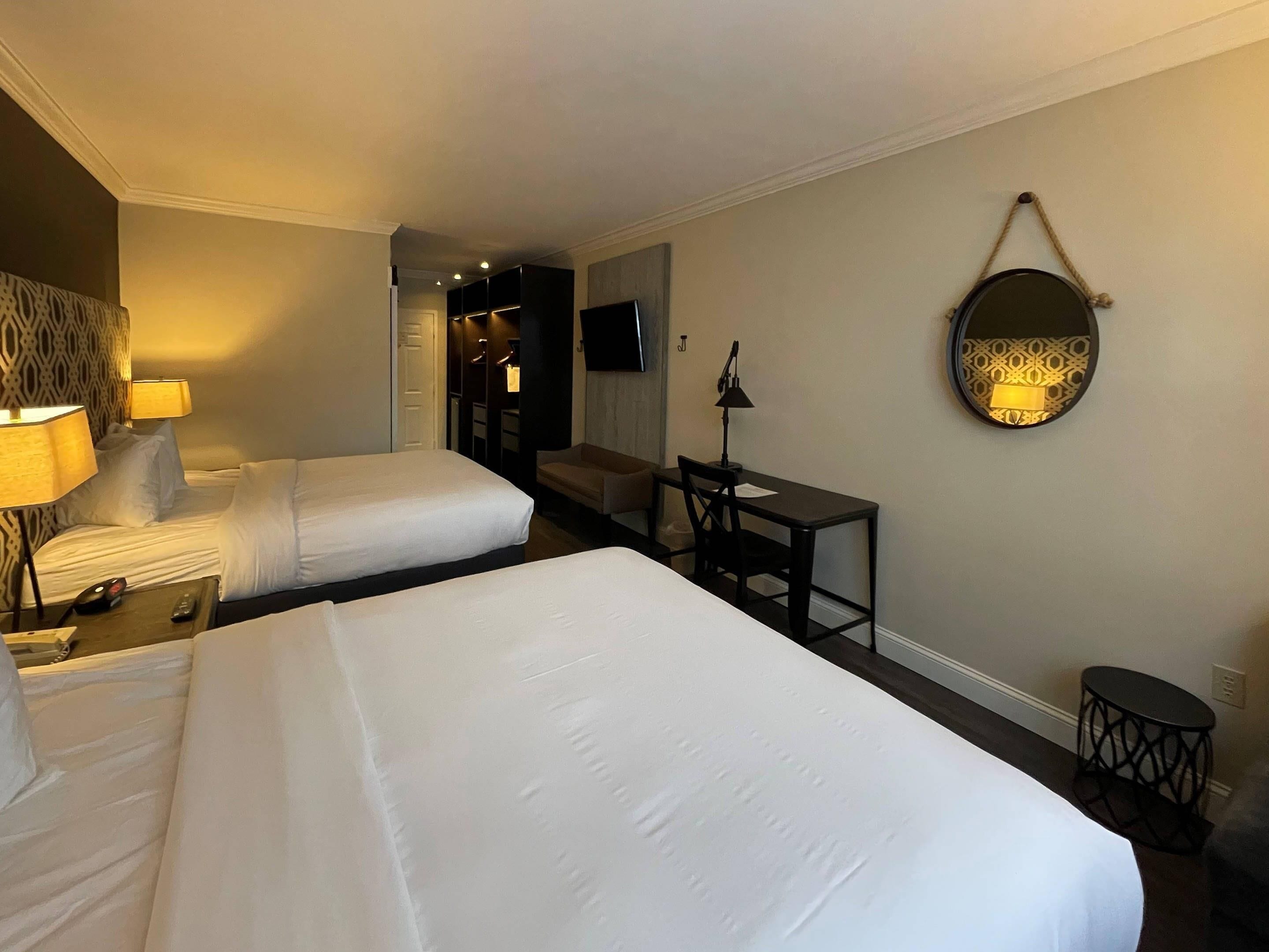 Habitación de hotel con dos camas, un escritorio, una silla, un televisor y un espejo en la pared.