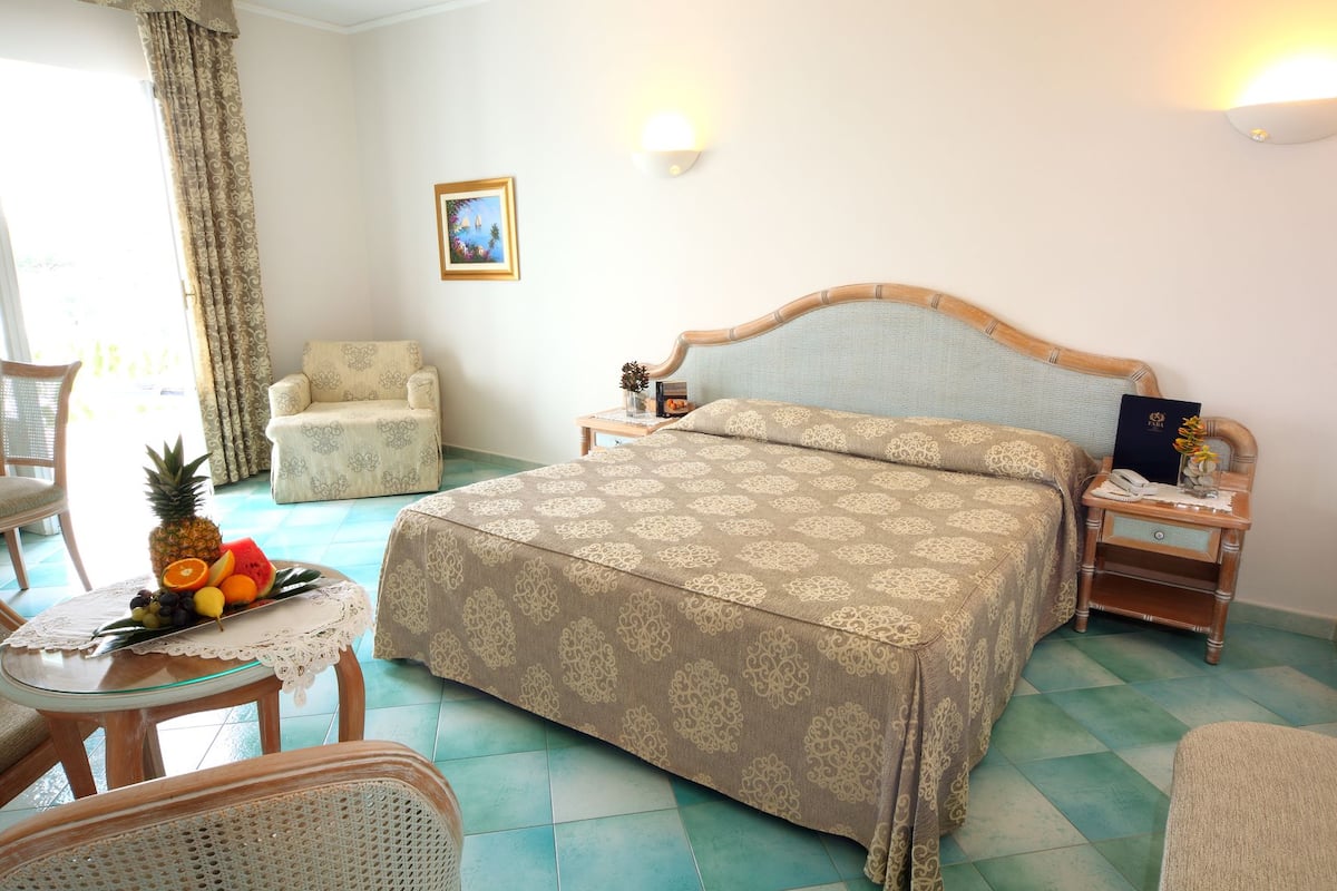A hotel room with a bed, a chair, a small table with fruit, and a nightstand with a book and a phone.