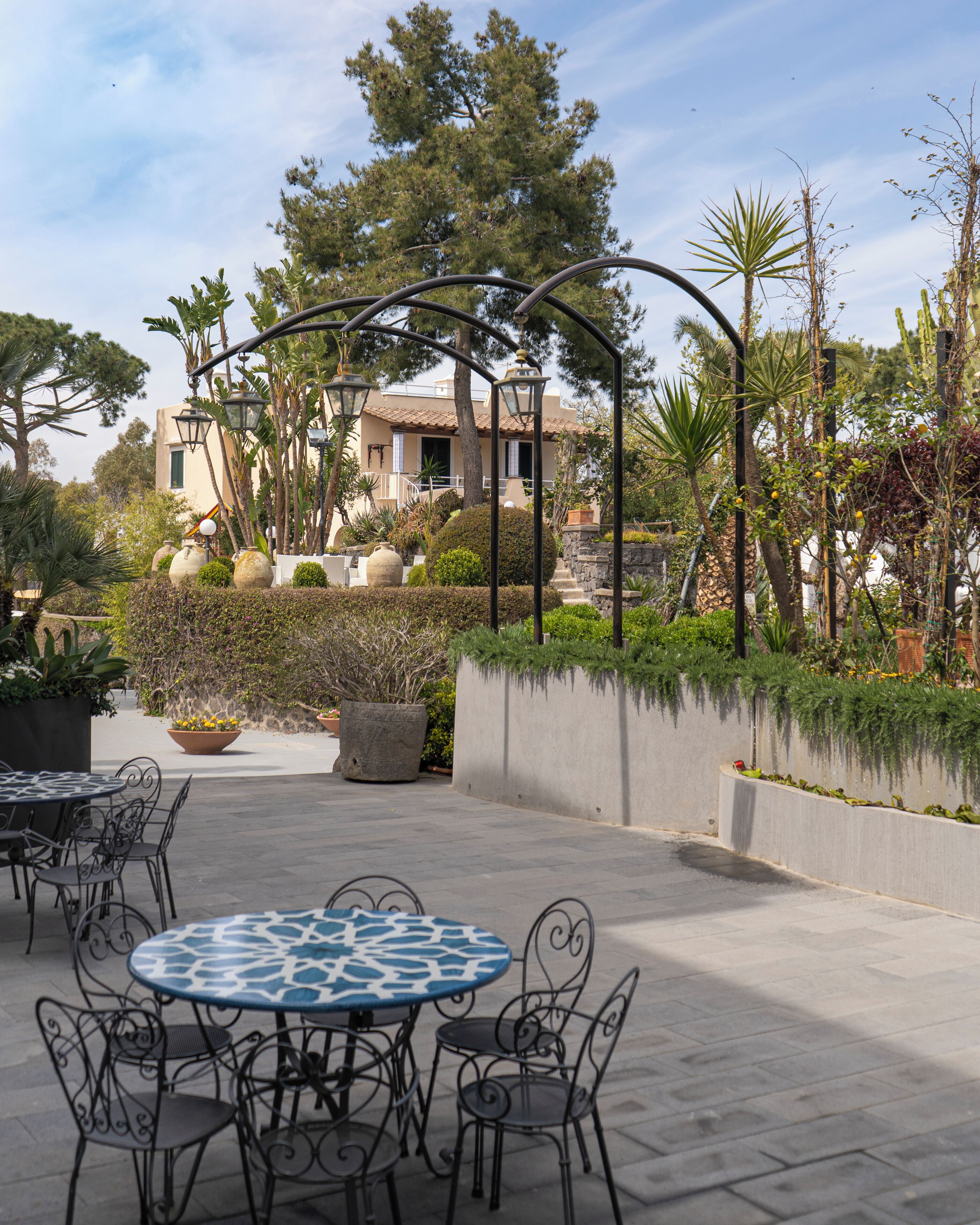 An outdoor patio area with round tables and chairs, surrounded by potted plants and a stone wall.