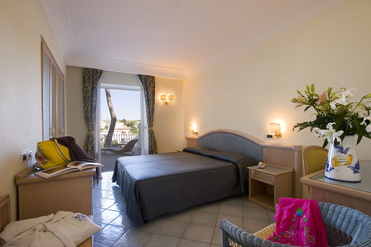 A hotel room with a bed, bedside tables, a chair, a small table with a vase of flowers, and a view of the outside.