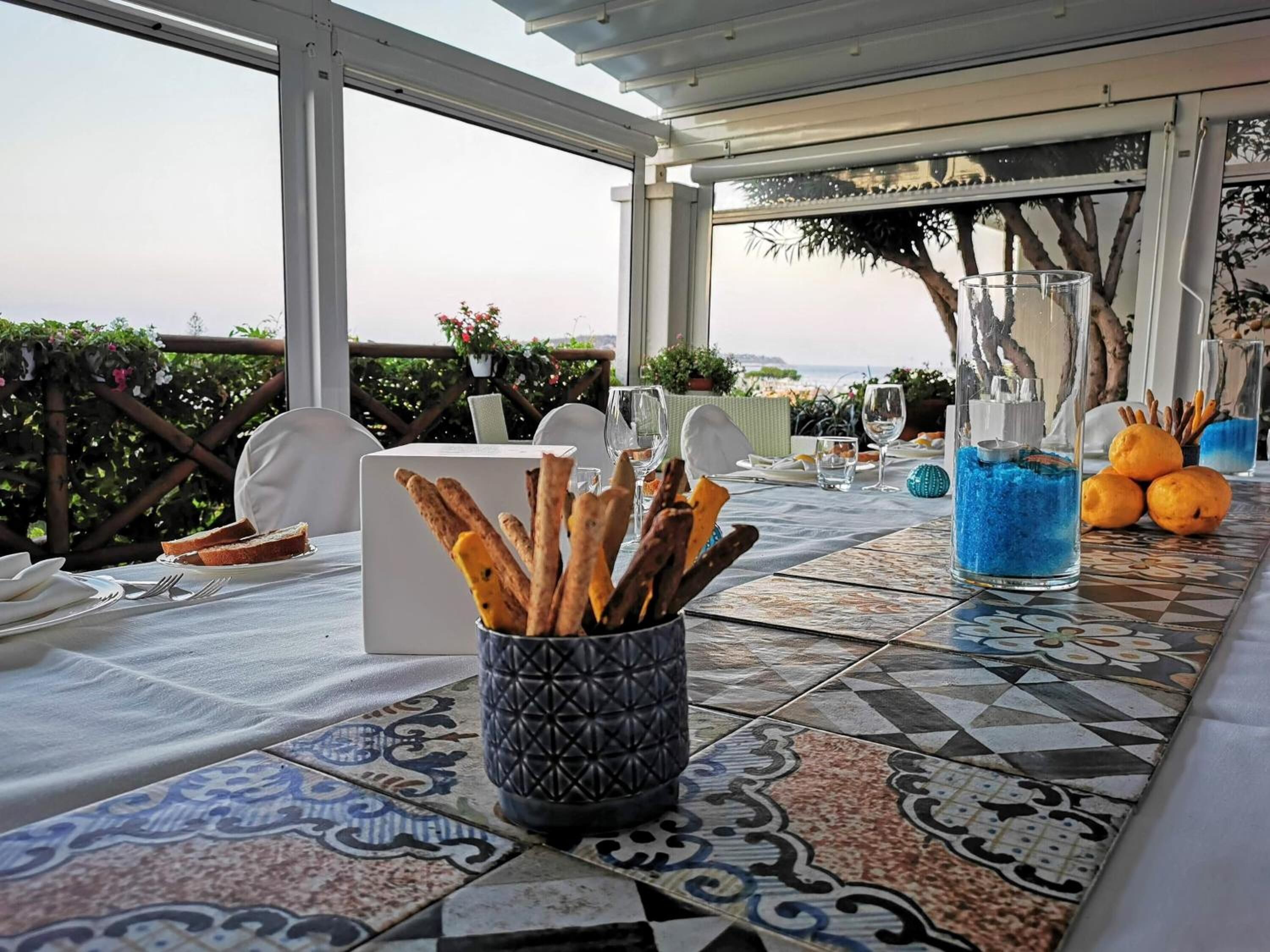 A table set for dining with a view of the sea, a glass vase with blue crystals, and a basket of breadsticks.