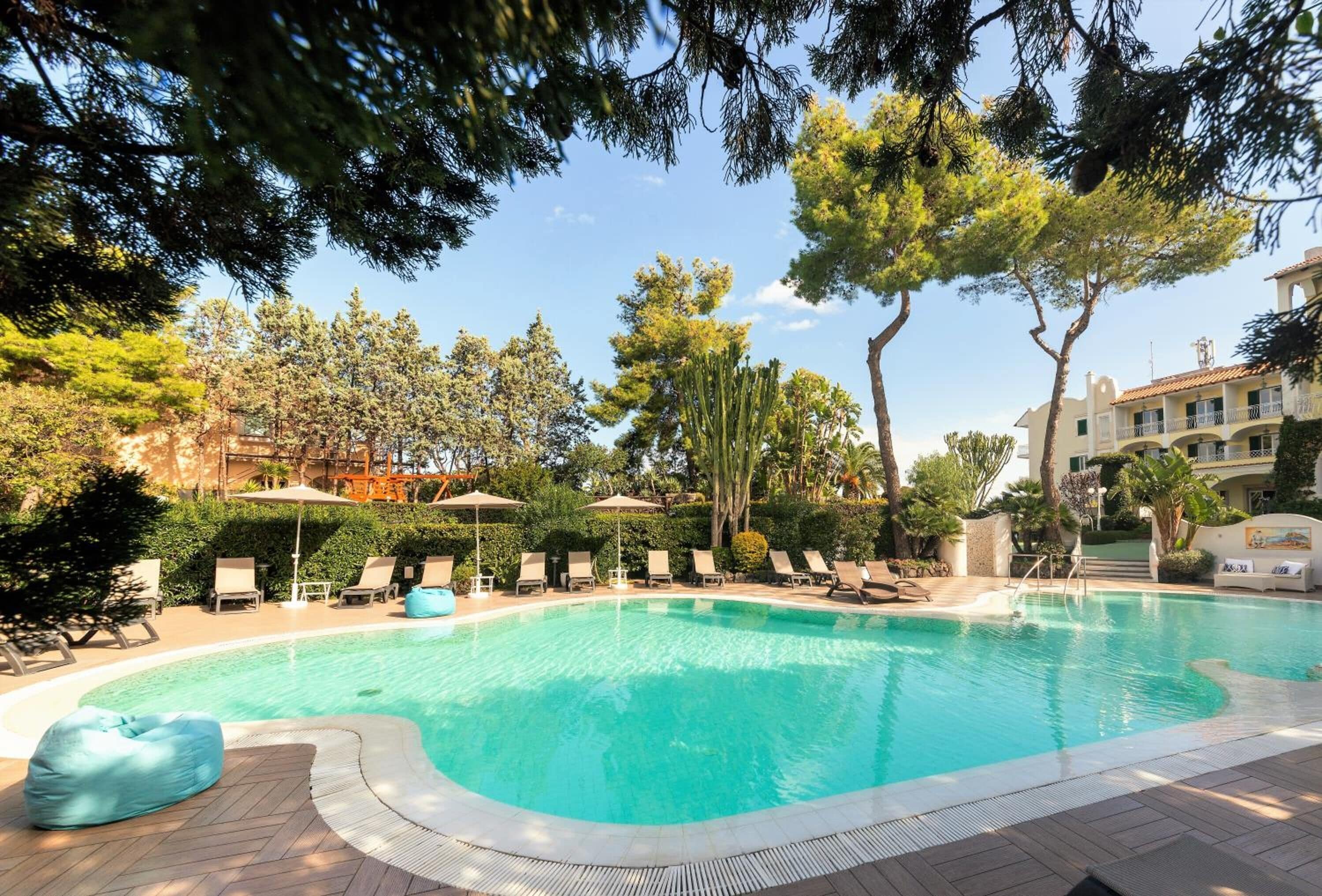 A swimming pool with lounge chairs and umbrellas, surrounded by trees and a building with balconies.