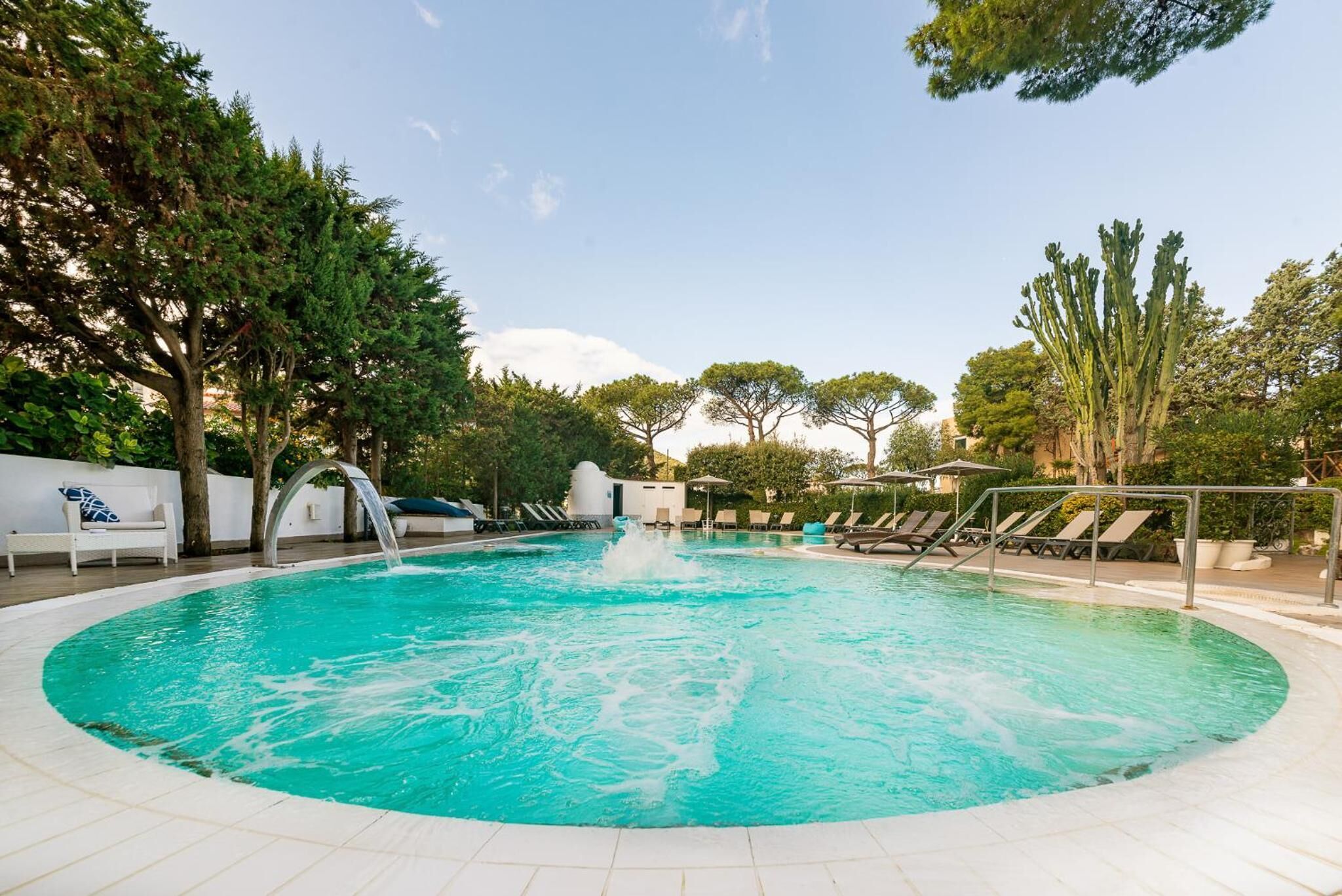A circular pool with a water feature, surrounded by lounge chairs and trees.
