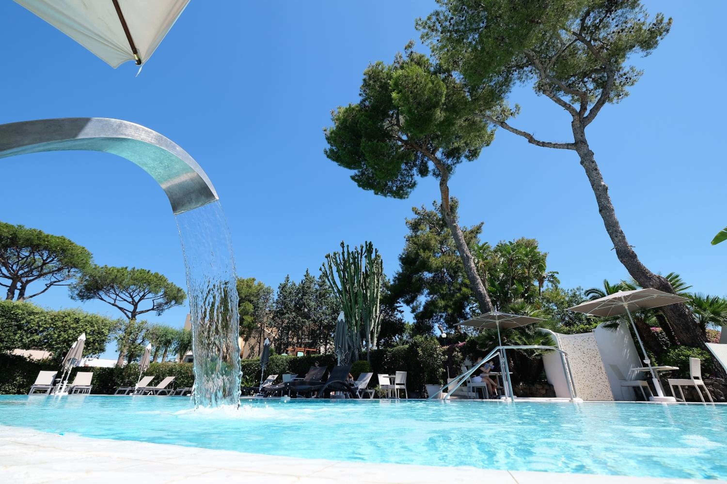 A swimming pool with a water feature, surrounded by lounge chairs and umbrellas, with trees in the background.
