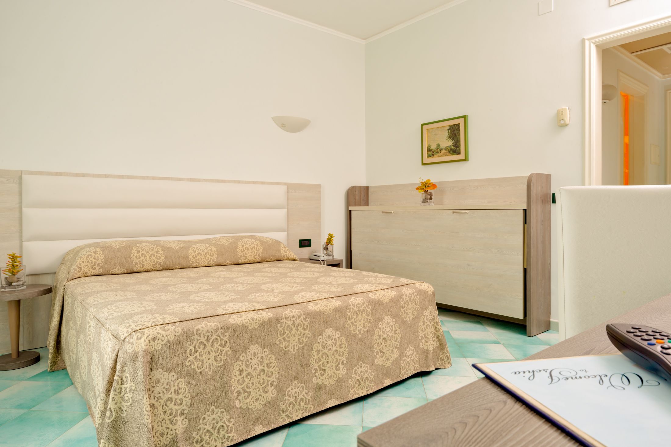 A bedroom with a bed, bedside tables, a nightstand, a dresser, a framed picture on the wall, and a remote control on a table.