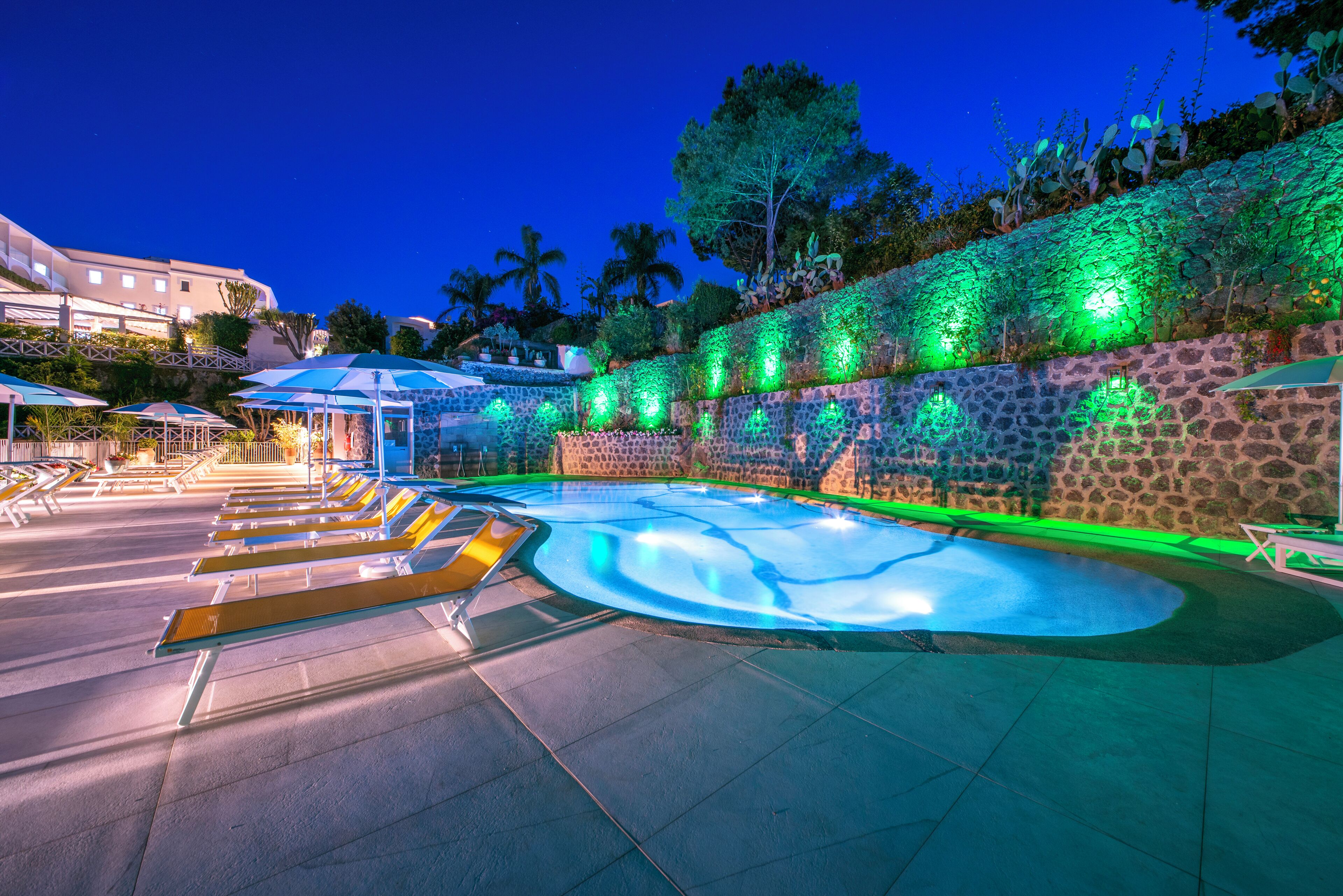 Zona de piscina con sillones de descanso y sombrillas, rodeada por un muro de piedra con follaje.