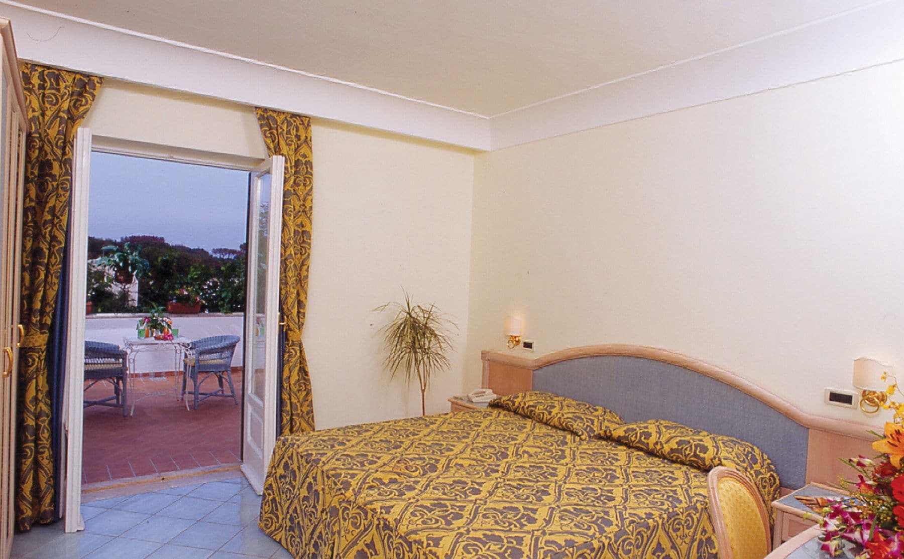 A bedroom with a bed, a nightstand, a lamp, a potted plant, and a view of an outdoor patio with a table and chairs.