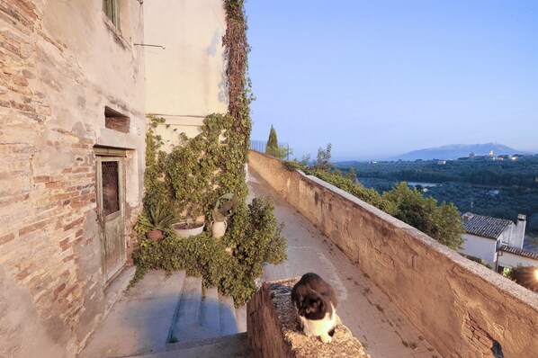 A narrow outdoor corridor with a stone wall on one side and a building on the other, leading to a view of a distant cityscape and mountains. - Castello Chiola (Loreto Aprutino)
