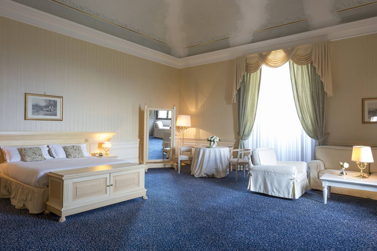 Habitación de hotel con una cama grande, una zona de estar con mesa y sillas, un escritorio con lámpara y un ventanal con cortinas.