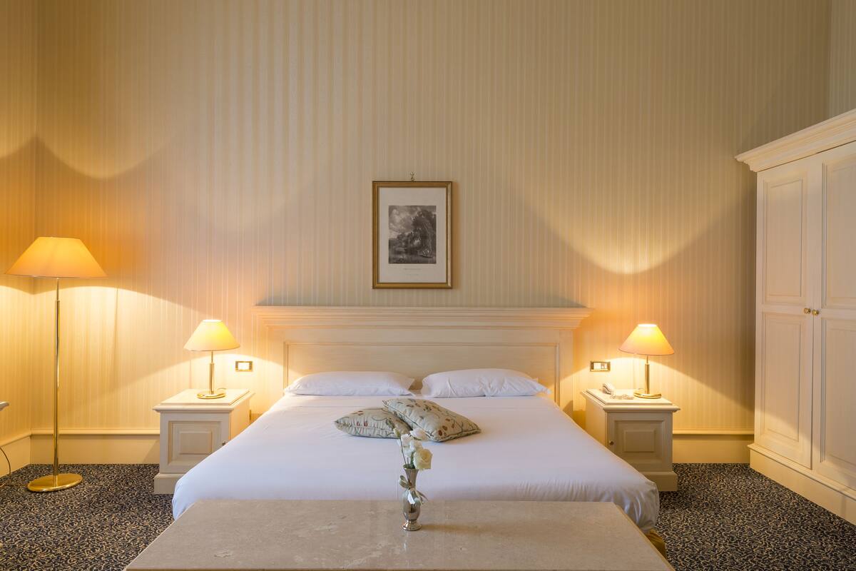 Habitación de hotel con una cama grande, dos lámparas de noche, un cuadro en la pared y un jarrón con flores sobre la mesita de noche.