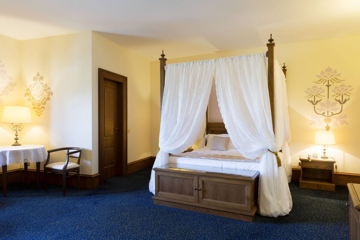 Una cama con dosel adornada con cortinas blancas, una mesita de noche de madera con una lámpara, una silla y una puerta en una habitación de hotel.