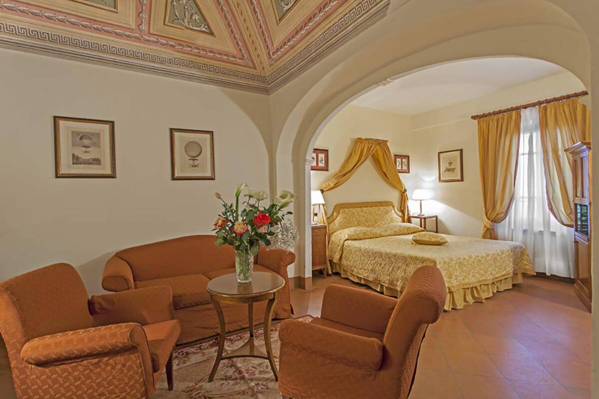 Habitación de hotel con una cama, dos sillones, una mesita con un florero y un techo decorativo.