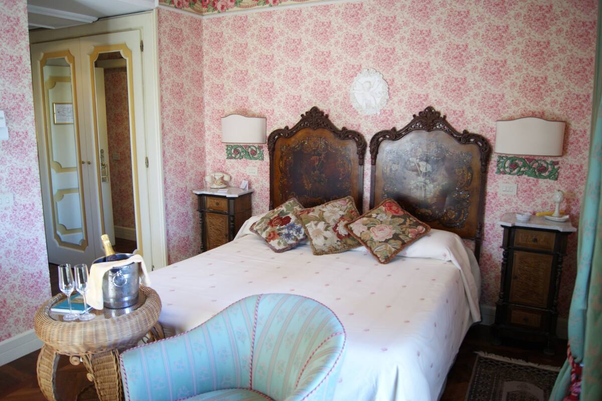 A bedroom with floral wallpaper, a wooden bed with floral pillows, a wicker chair, and a bedside table with a lamp.