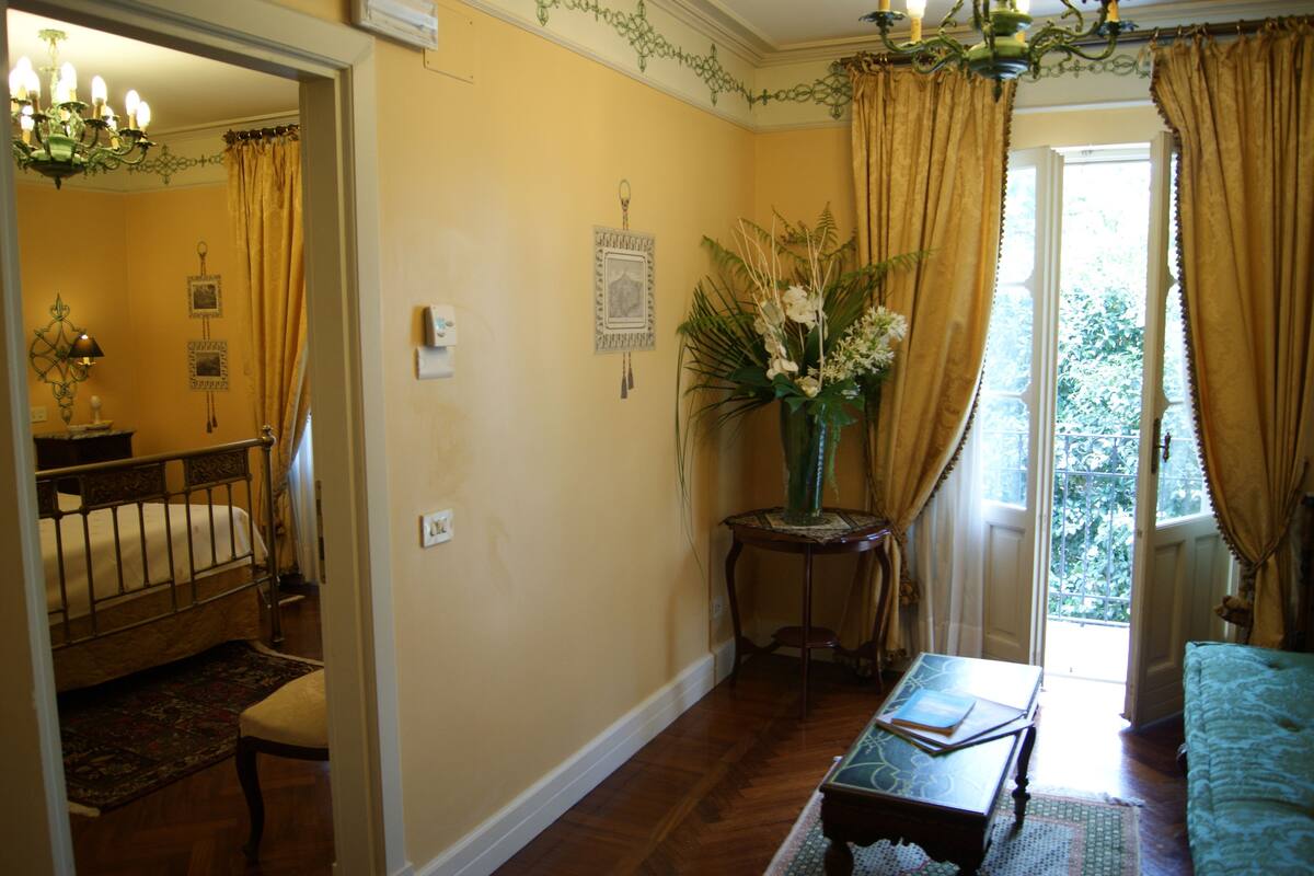 A room with a bed, a small table with a vase of flowers, a chair, and a window with curtains.