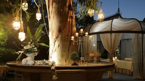 A well-lit outdoor area with a large tree, a decorative fountain, hanging lanterns, and a covered seating area. - Hotel Villa Paradiso dell'Etna (San Giovanni la Punta)