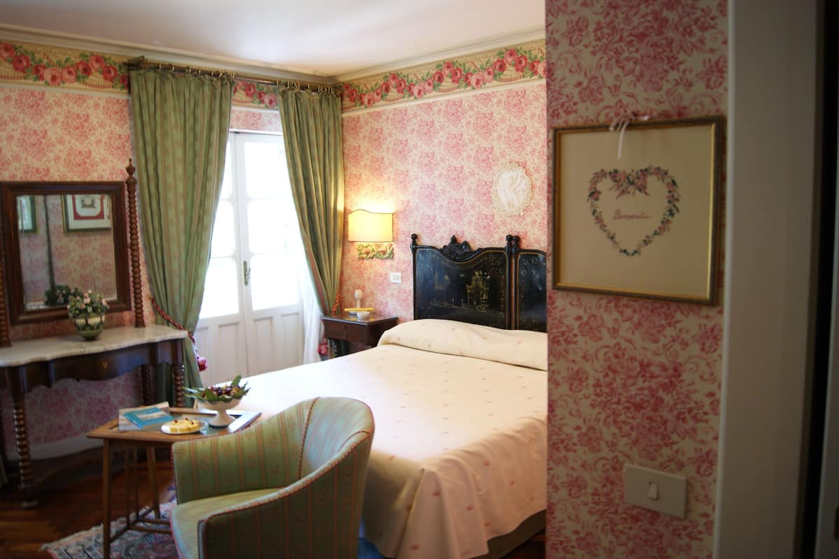 A bedroom with floral wallpaper, a bed with a heart-shaped decoration, a bedside table with a lamp, and a chair.