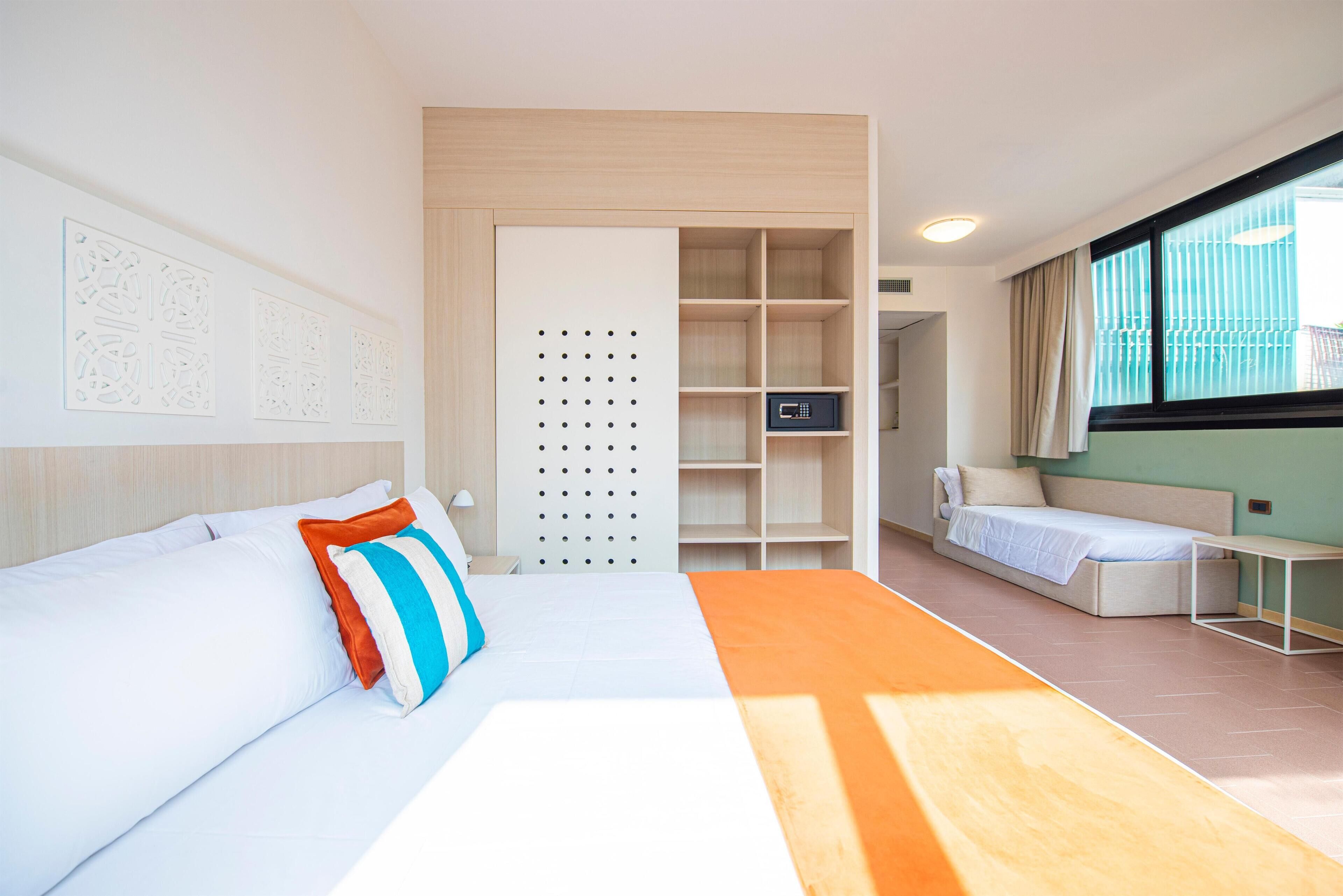 A modern hotel room with a bed, a desk, a wardrobe, and a view of the outside.